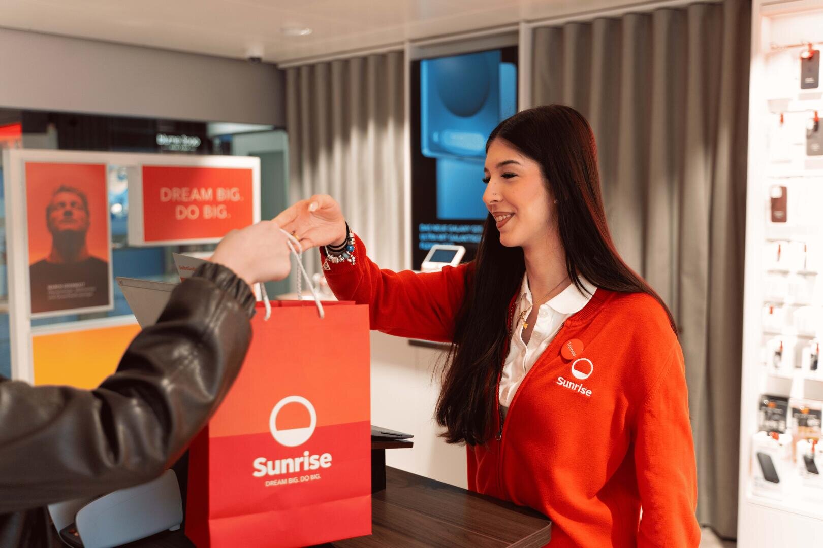 Eine lächelnde Frau in einer roten „Sunrise“-Uniform reicht einem Kunden in einem modernen Einzelhandelsgeschäft über den Tresen hinweg eine Einkaufstasche mit dem Firmenlogo.