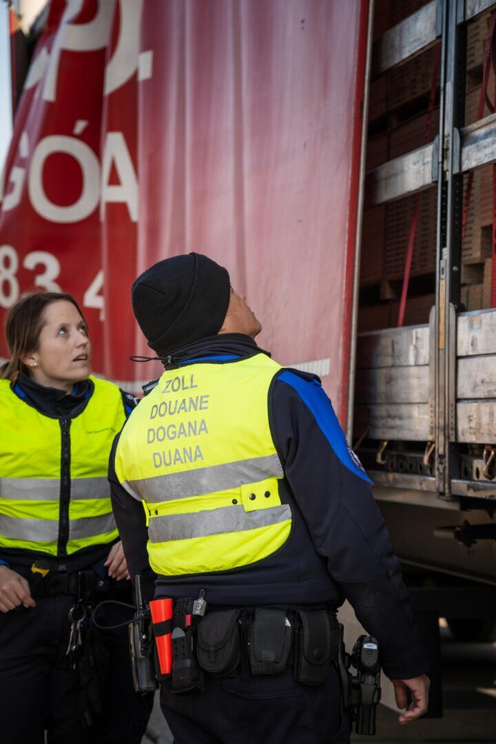 Zwei Zollbeamte in gelben Westen inspizieren die offene Seite eines Lastwagens. Ein Beamter blickt in den Lastwagen, der andere mit besorgtem Blick. Die Seite des Lastwagens ist rot mit weißer Schrift.