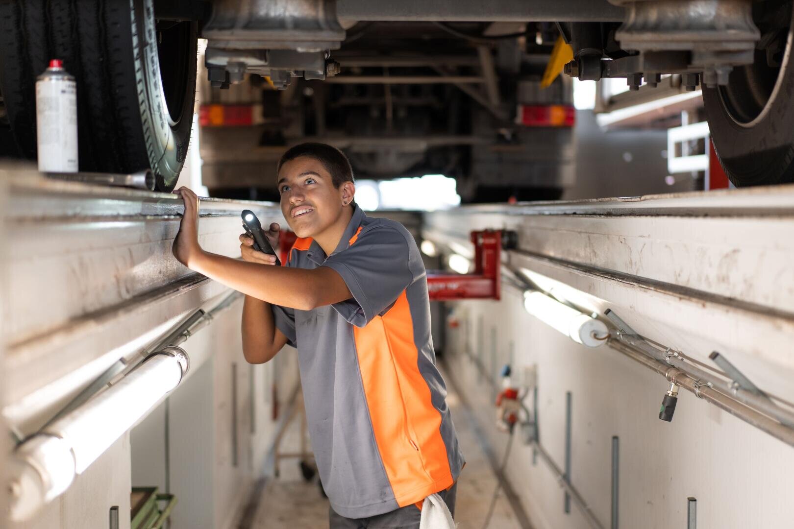 Ein Mechaniker in einer grau-orangefarbenen Arbeitskleidung lächelt, während er mit einer Taschenlampe in der Hand in einer hell beleuchteten Werkstatt die Unterseite eines Fahrzeugs untersucht.
