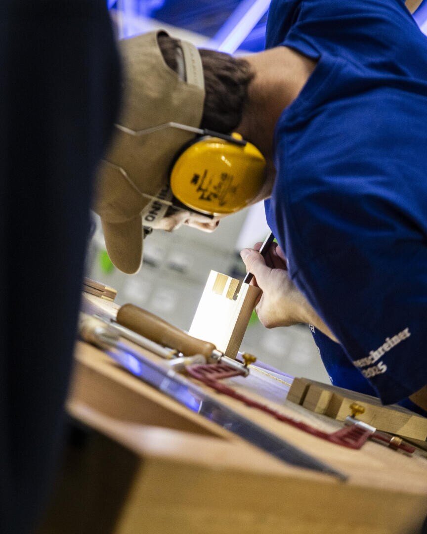 Eine Person, die einen Gehörschutz und eine Mütze trägt, arbeitet an einem Holzbearbeitungsprojekt, wobei sie einen Meißel und einen Hammer auf einem Stück Holz an einer Werkbank verwendet. In der Nähe sind Werkzeuge und Klemmen zu sehen.