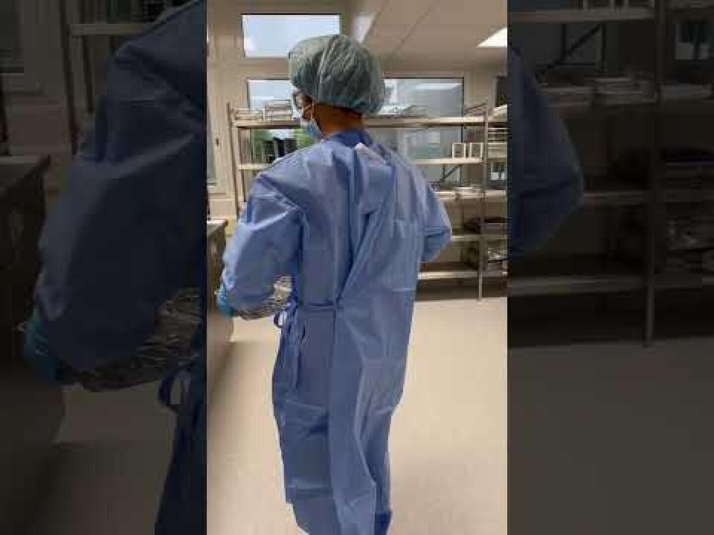 Eine Person, die blaue OP-Kleidung, einen Haarschutz und eine Maske trägt, geht durch eine sterile medizinische Einrichtung, in der im Hintergrund Regale und Geräte zu sehen sind.