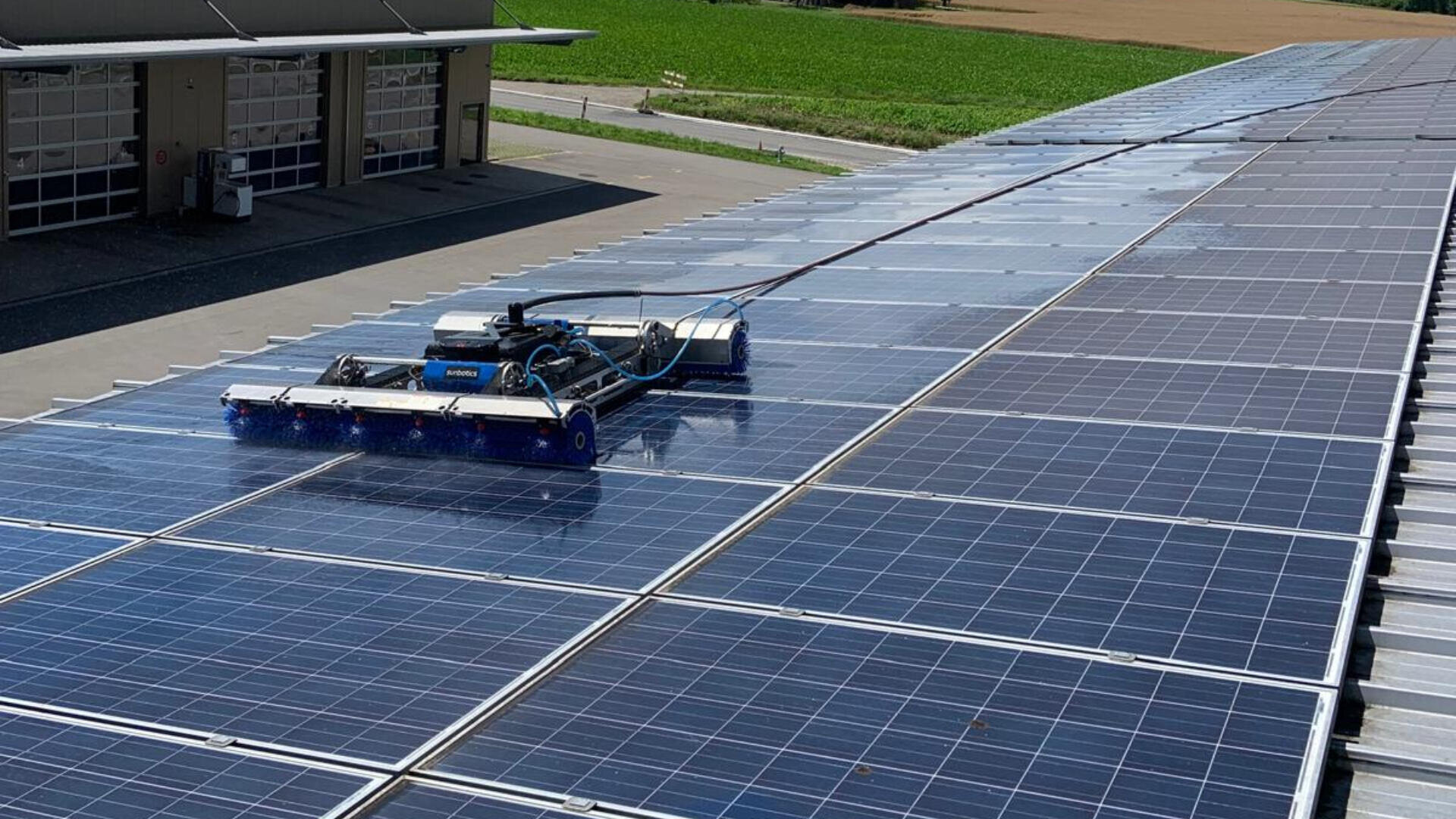 Ein Roboter fährt über große Reihen von Solarmodulen auf einem Dach und reinigt die Oberfläche. Im Hintergrund sind ein Gebäude und grüne Felder zu sehen.