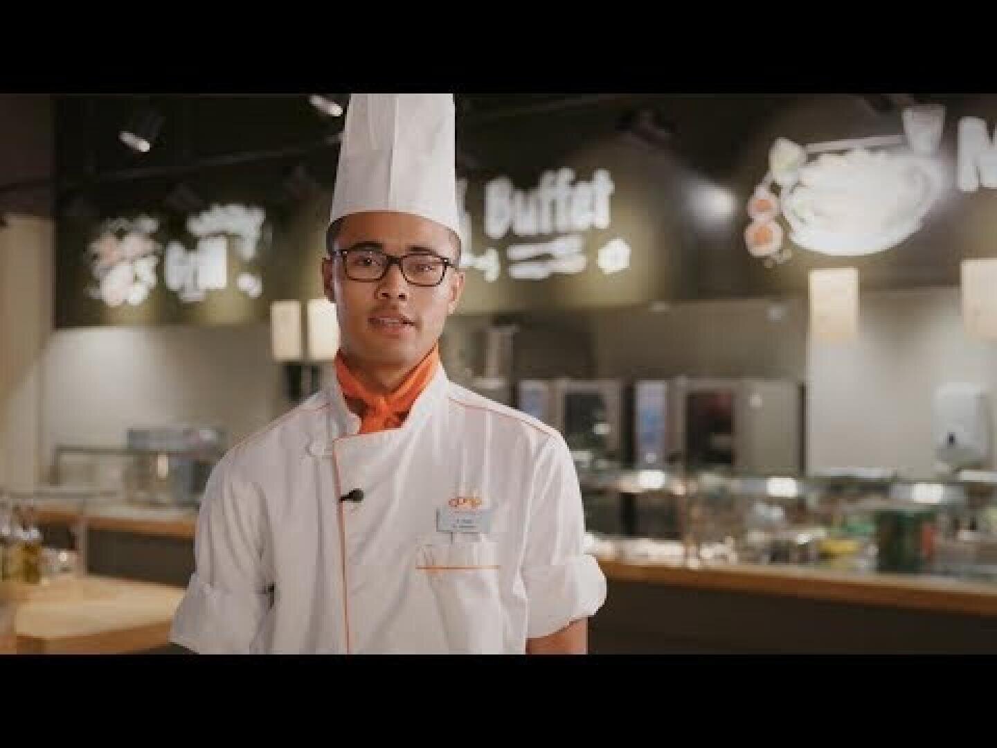Ein Koch mit einer hohen weißen Kochmütze, einer Brille und einer weißen Kochuniform mit orangefarbenem Halstuch steht in einer Restaurantküche, im Hintergrund sind Buffetschilder zu sehen.
