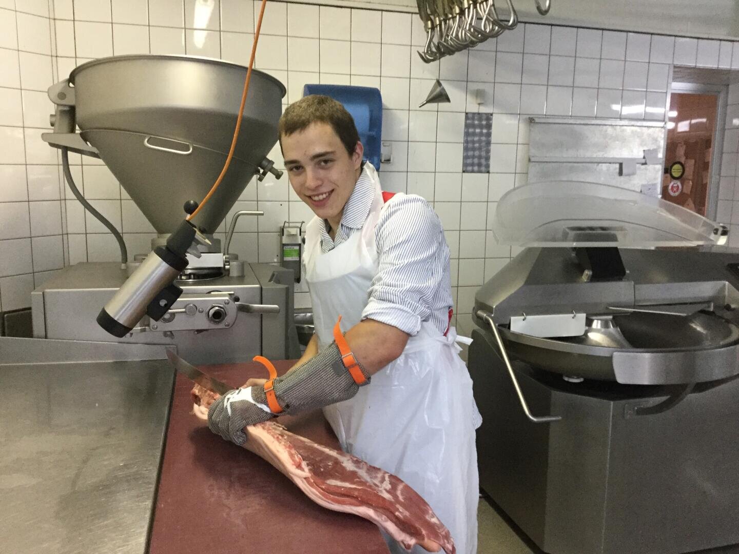 Ein junger Mann mit weißer Schürze und Schutzhandschuhen lächelt, während er ein großes Stück rohes Fleisch in einer gekachelten Großküche oder Metzgerei zubereitet.