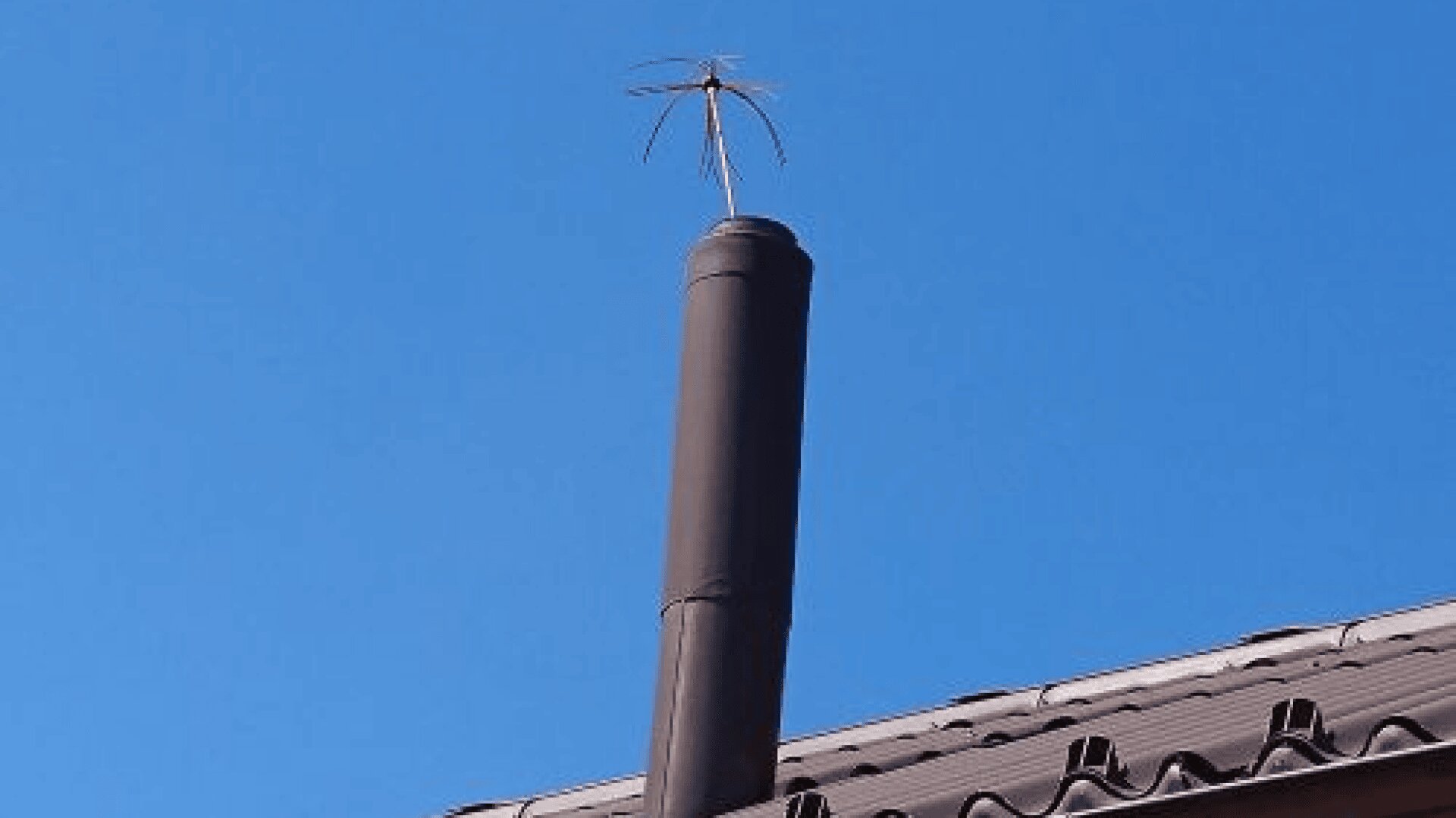 Ein metallenes Schornsteinrohr ragt aus einem Ziegeldach gegen einen klaren blauen Himmel, an dessen Spitze eine kleine kreisförmige Drahtantenne oder ein Schutz angebracht ist.