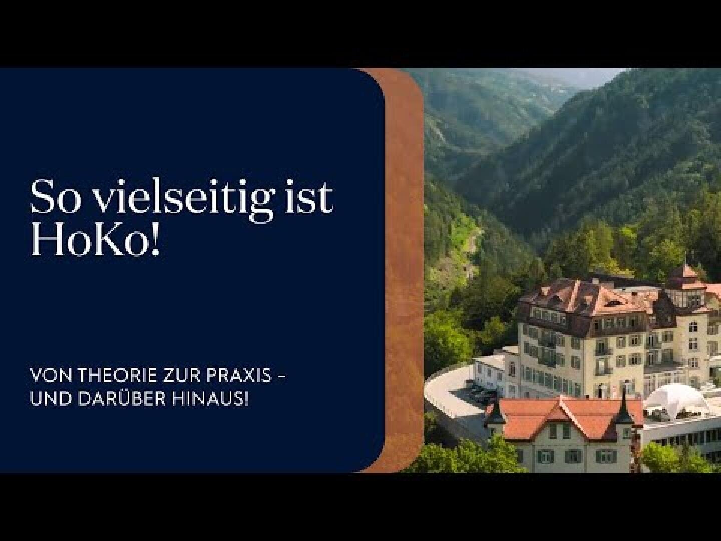 Ein großes, elegantes Gebäude mit roten Dächern liegt inmitten von grünen, bewaldeten Hügeln unter einem klaren Himmel. Der Text auf einer dunkelblauen Tafel lautet: So vielseitig ist HoKo! Von der Theorie zur Praxis - und darüber hinaus!.