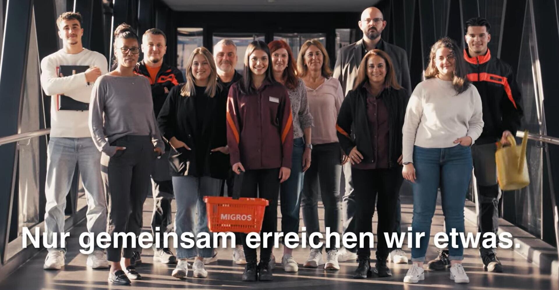 Eine bunt gemischte Gruppe von zwölf Personen steht lächelnd in einem sonnendurchfluteten Korridor zusammen. Eine Frau hält einen orangefarbenen Migros-Korb in der Hand. Der deutsche Text lautet: Nur gemeinsam erreichen wir etwas.