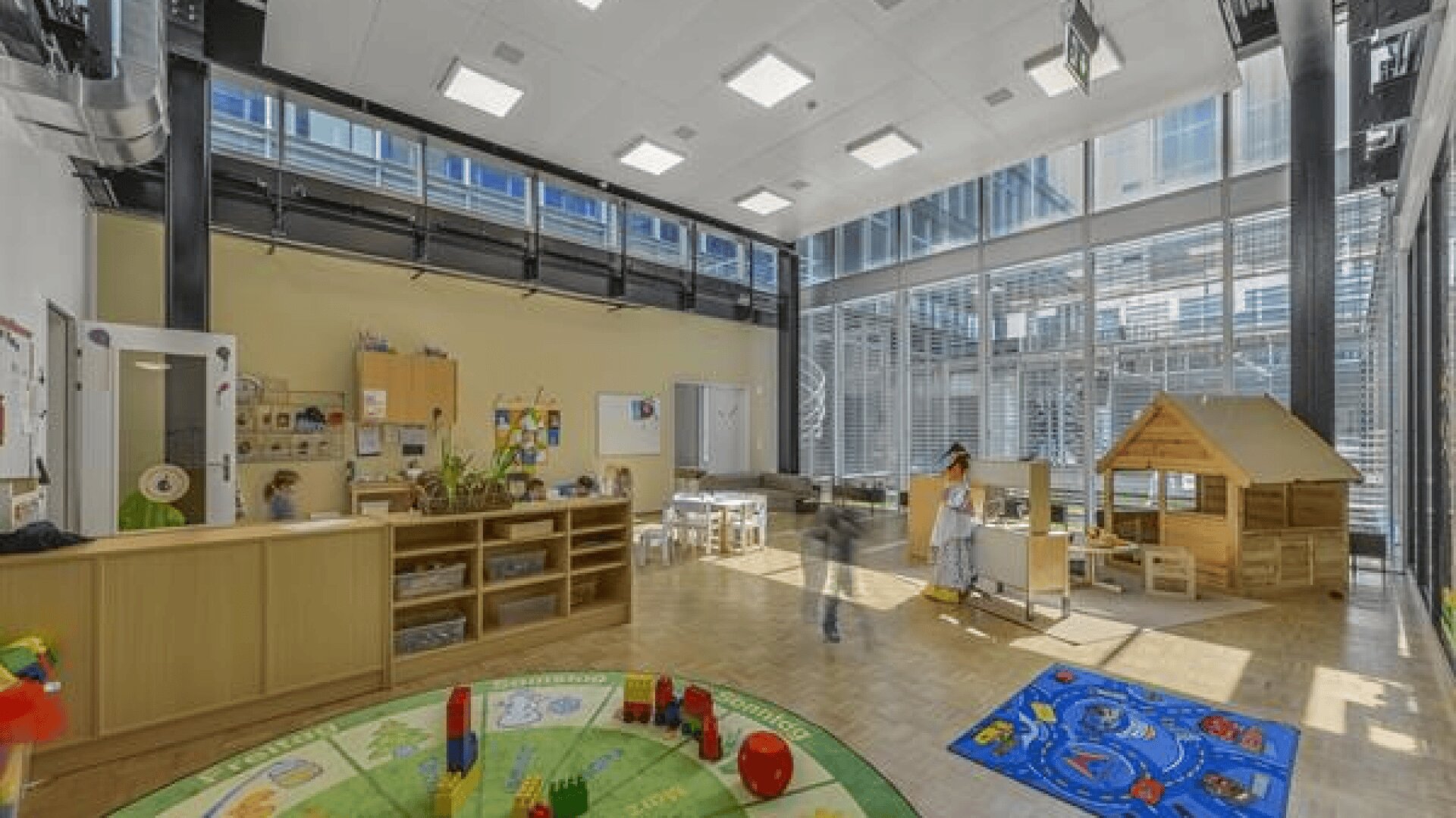 Ein geräumiges, helles Klassenzimmer mit großen Fenstern, Regalen mit Spielzeug, einem Spielhaus, einer Leseecke und Kindern, die sich mit verschiedenen Aktivitäten beschäftigen. Der Raum ist mit bunten Matten und pädagogischer Dekoration ausgestattet.