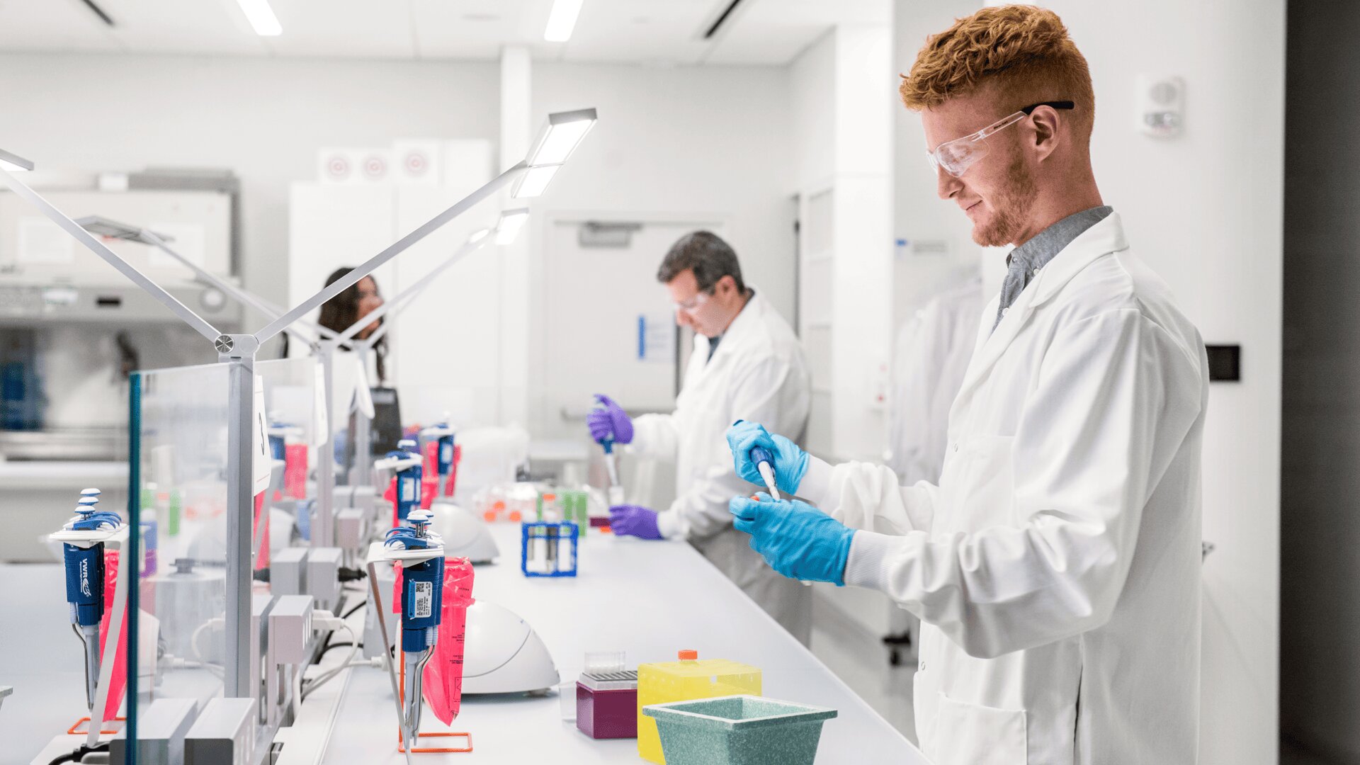 Zwei Wissenschaftler in weißen Laborkitteln und blauen Handschuhen arbeiten an einem Labortisch mit wissenschaftlichen Geräten, Fläschchen und Röhrchen. Das Labor ist sauber, hell und modern, mit weißen Wänden und Schutzbrillen, die von den Mitarbeitern getragen werden.