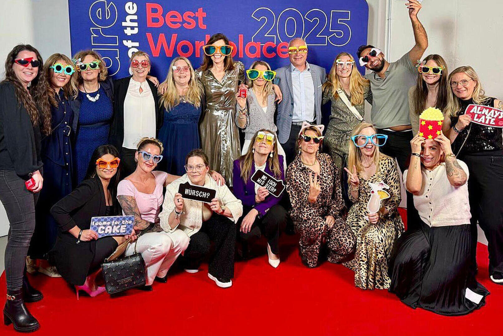Eine Gruppe von Menschen posiert auf einem roten Teppich vor einer Kulisse der Best Workplaces 2025, trägt bunte Brillen und hält verspielte Requisiten in der Hand, lächelt und feiert gemeinsam.