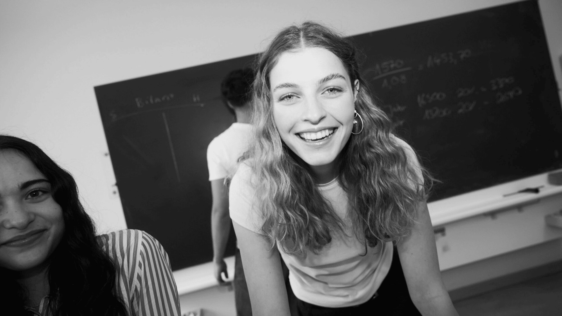 Eine junge Frau mit welligem Haar lächelt in einem Klassenzimmer strahlend in die Kamera. Neben ihr steht ein weiterer Schüler, im Hintergrund ist eine Tafel zu sehen. Das Foto ist schwarz-weiß.