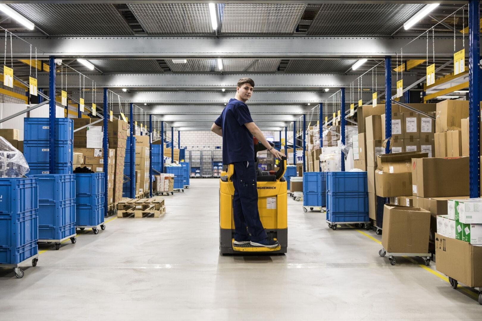 Ein Lagerarbeiter in blauer Uniform bedient einen gelben Hubwagen und transportiert Kartons durch einen breiten Gang, der von Regalen und blauen Lagerbehältern gesäumt ist, in einem hell beleuchteten Lager.