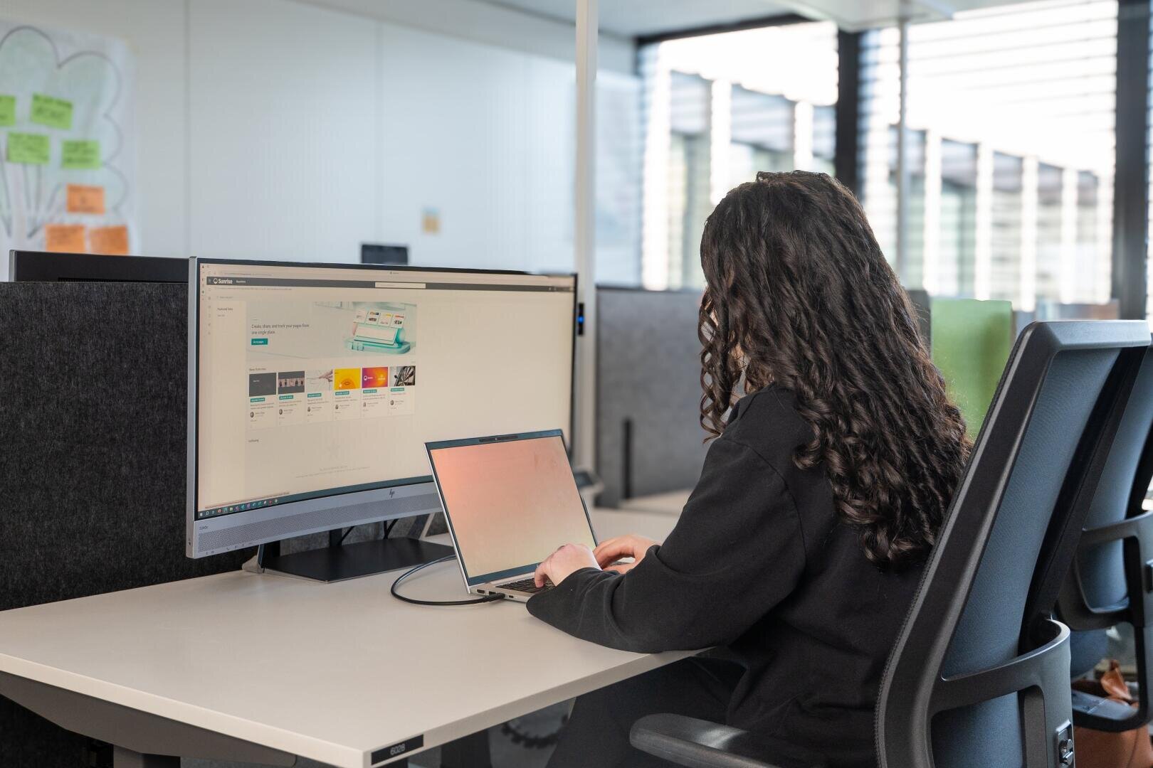Eine Person mit langen lockigen Haaren arbeitet an einem Schreibtisch mit einem Laptop, der an einen großen Monitor in einem modernen Büro mit abgetrennten Arbeitsbereichen und natürlichem Licht angeschlossen ist.