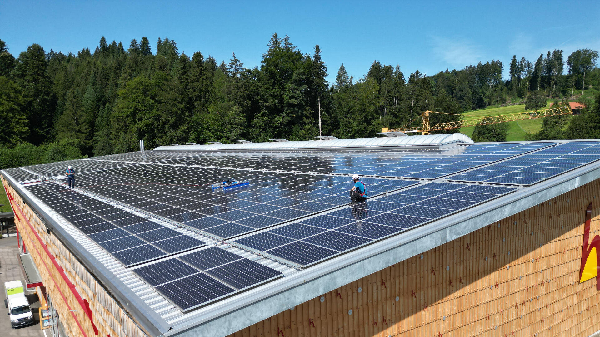 Arbeiter in Sicherheitsausrüstung installieren oder warten Solarmodule auf dem Dach eines großen Holzgebäudes, umgeben von Bäumen und einem klaren blauen Himmel.