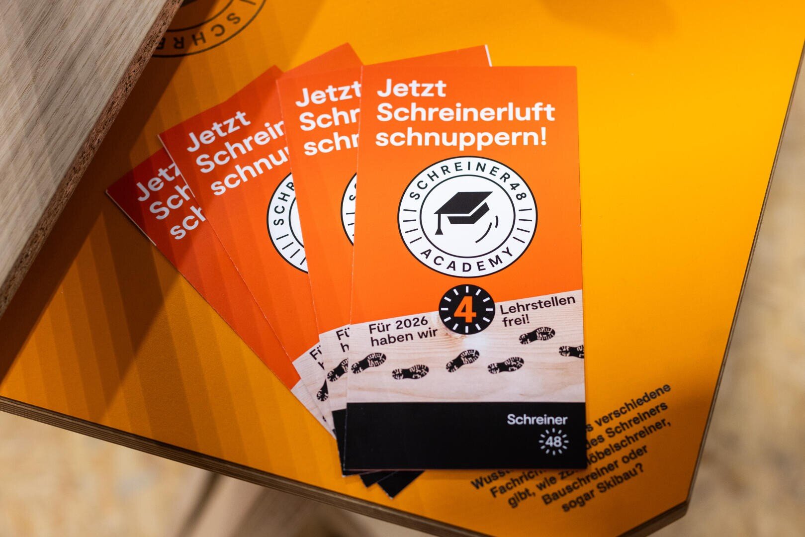 Vier orangefarbene Flugblätter mit weißem und schwarzem Text in deutscher Sprache, die für eine Akademie werben. Der Text lautet "Jetzt Schreinerluft schnuppern!" mit einem Logo und Angaben zu vier freien Ausbildungsplätzen im Jahr 2026.
