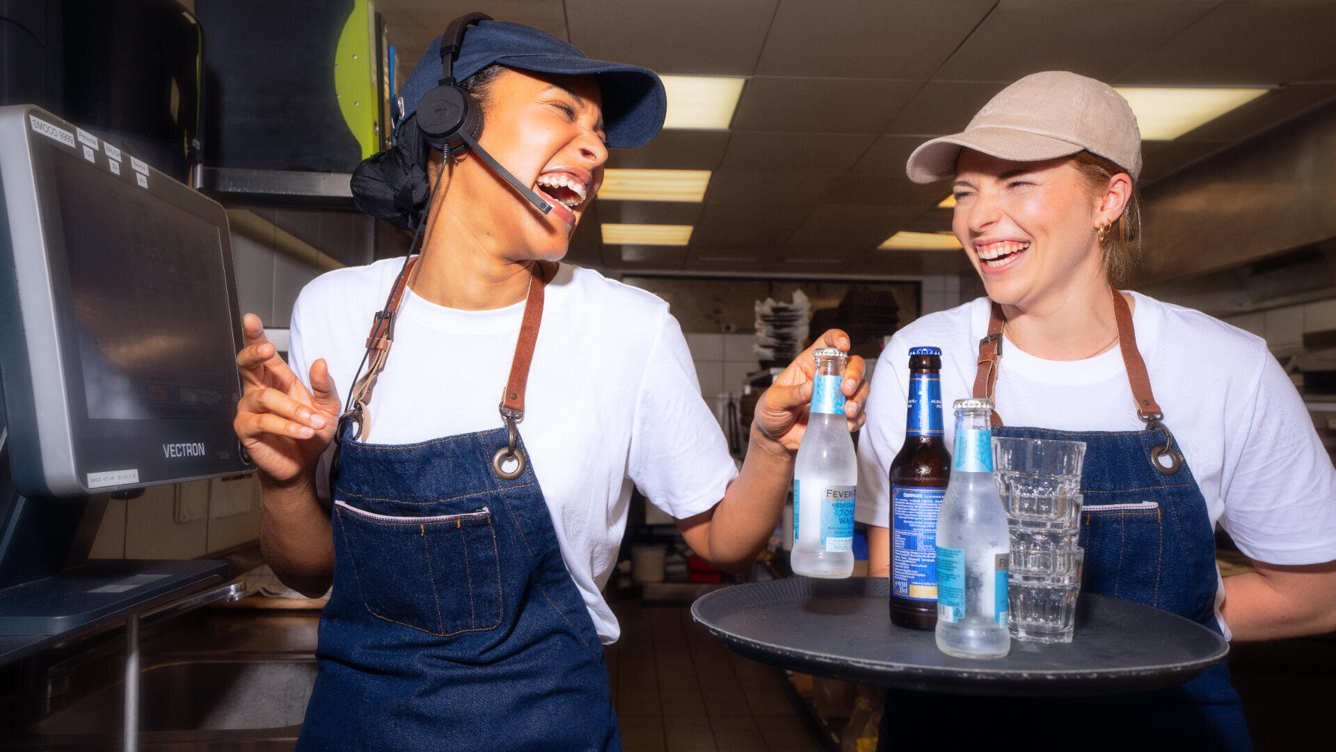 Zwei Restaurantangestellte in blauen Schürzen und Hüten lachen zusammen in einer Küche. Die eine hält ein Tablett mit Getränkeflaschen und Gläsern, die andere trägt ein Headset und gestikuliert mit der Hand. Beide wirken fröhlich und glücklich.