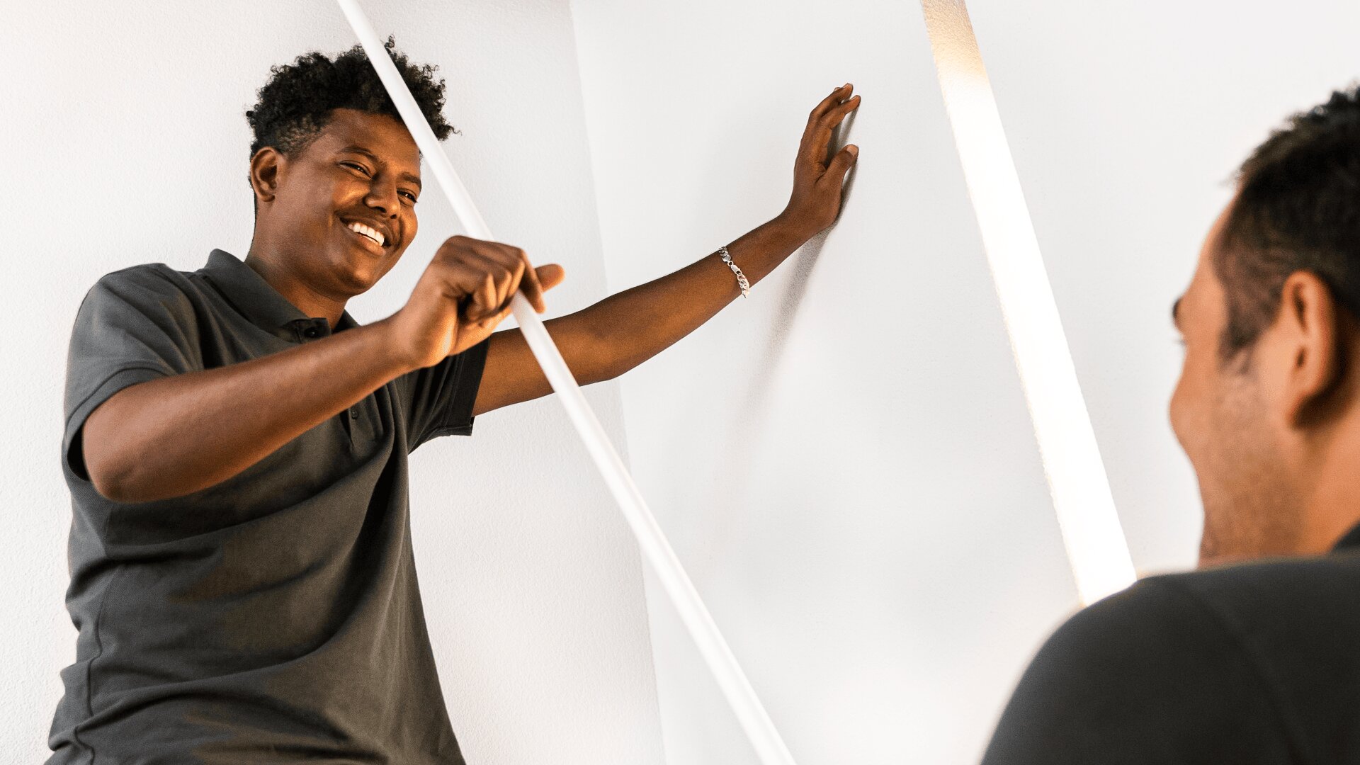 Zwei Personen stehen in einem Haus und lächeln sich an; die eine lehnt an einer weißen Wand und hält einen langen weißen Gegenstand, während die andere Person teilweise im Vordergrund zu sehen ist.