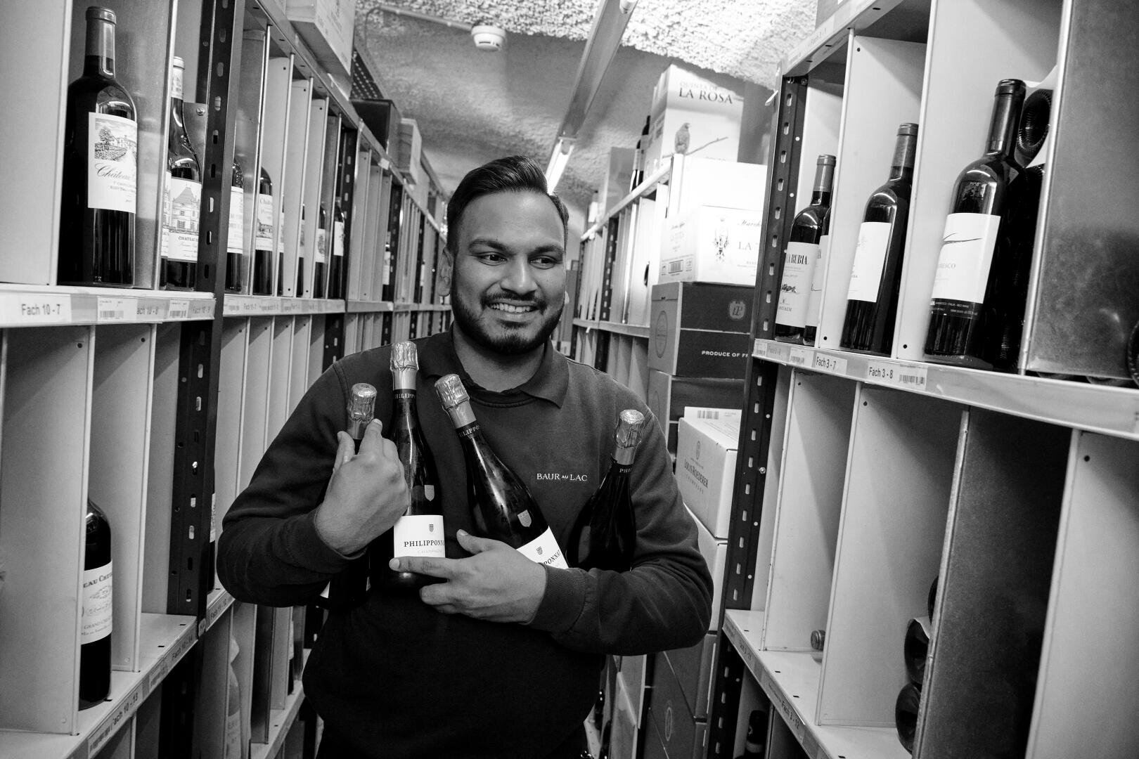 Ein Mann steht zwischen mit Weinflaschen gefüllten Regalen und hält lächelnd mehrere Weinflaschen in seinen Armen. Das Bild ist in schwarz-weiß gehalten.
