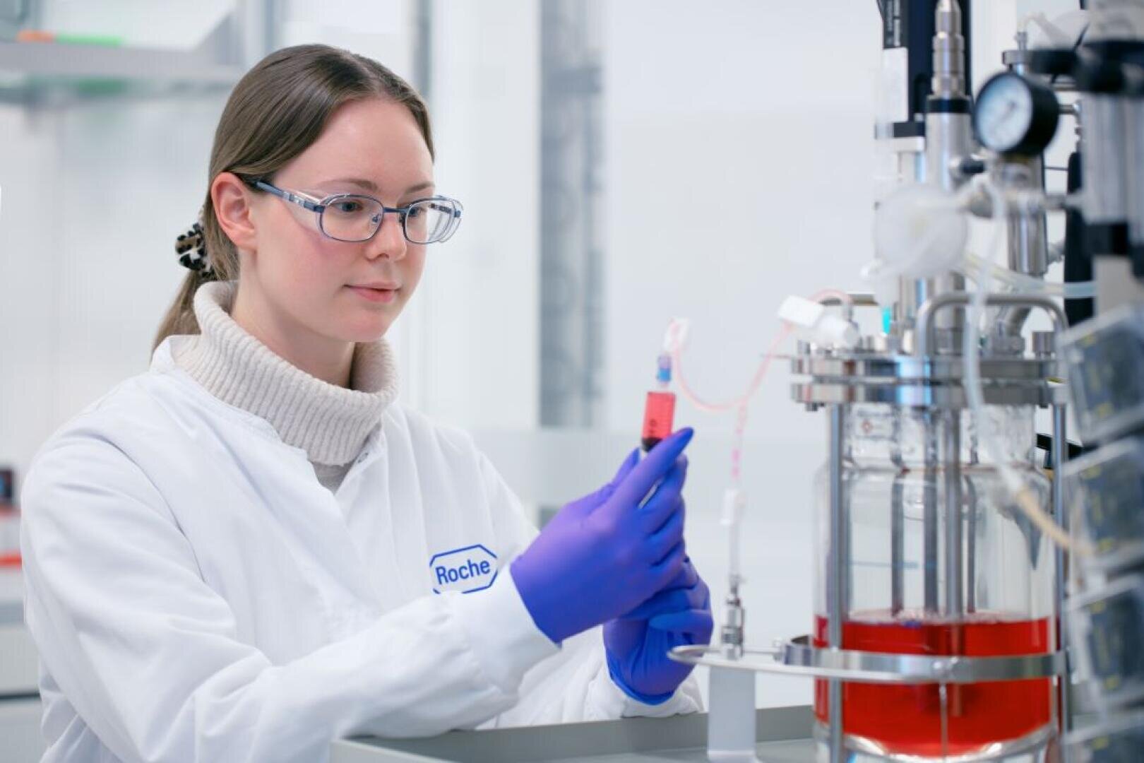Ein Wissenschaftler, der eine Brille, Handschuhe und einen weißen Laborkittel mit der Aufschrift Roche trägt, arbeitet in einem Labor, hält eine Pipette in der Hand und hantiert mit einem mit roter Flüssigkeit gefüllten Fläschchen in der Nähe wissenschaftlicher Geräte.