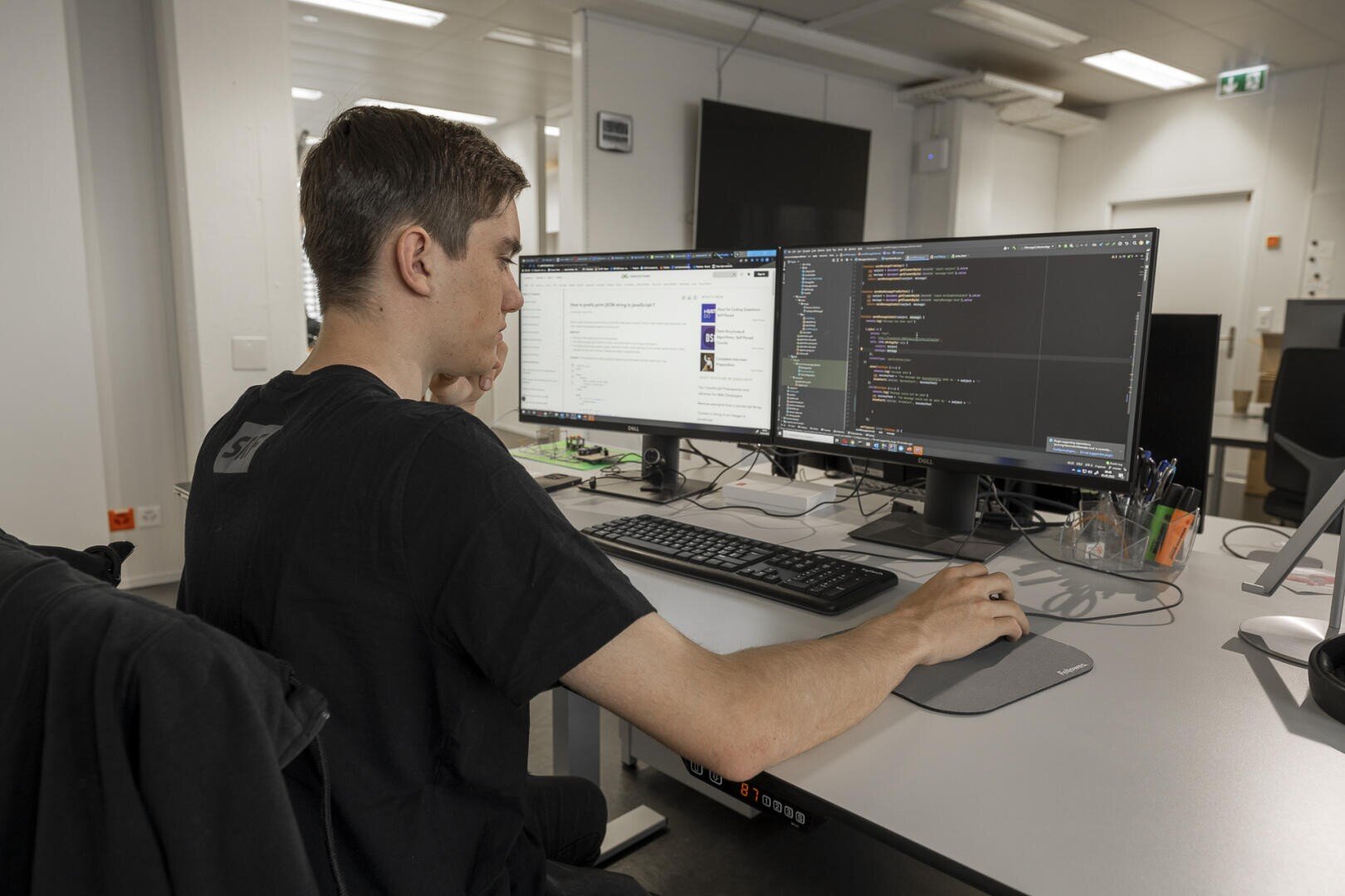 Eine Person sitzt an einem Schreibtisch und arbeitet an zwei Computermonitoren, von denen einer einen Code und der andere eine Webseite anzeigt. Auf dem Schreibtisch befinden sich eine Tastatur, eine Maus, ein Stifthalter und Büromaterial in einer modernen Büroumgebung.