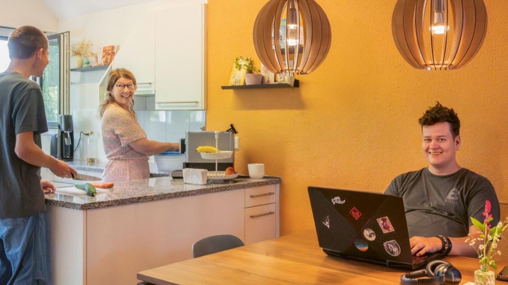Drei Personen befinden sich in einer gemütlichen Küche mit warmen gelben Wänden. Zwei von ihnen bereiten am Tresen Essen zu, während eine Person am Tisch sitzt, lächelt und an einem mit Aufklebern verzierten Laptop arbeitet.