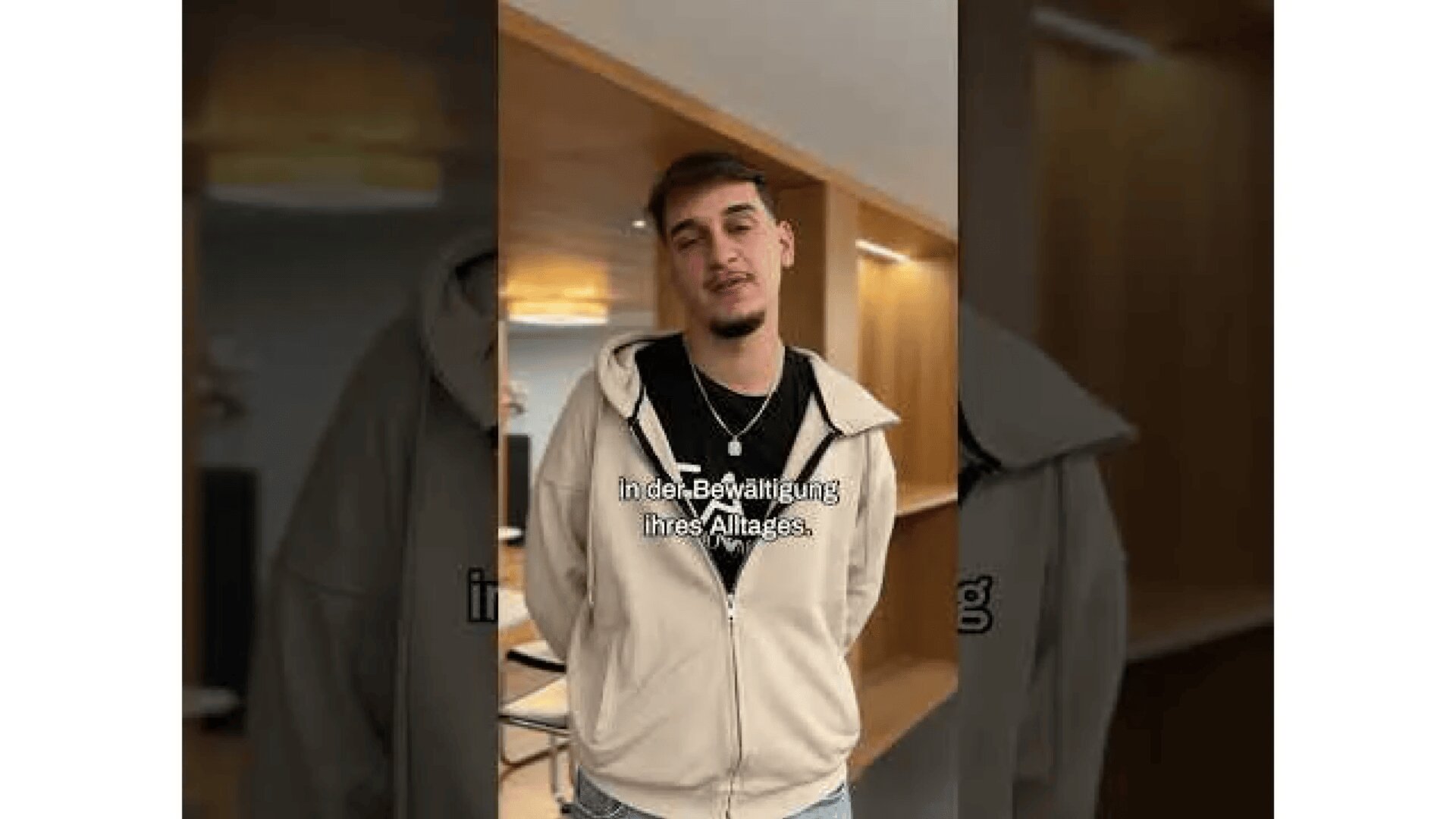 Ein junger Mann mit kurzen Haaren und gestutztem Bart steht in einem Haus und trägt einen beigen Kapuzenpullover und ein schwarzes T-Shirt. Der deutsche Text in der Bewältigung ihres Alltags. erscheint auf dem Bild. Der Hintergrund zeigt einen modernen Raum mit Holzvertäfelung.
