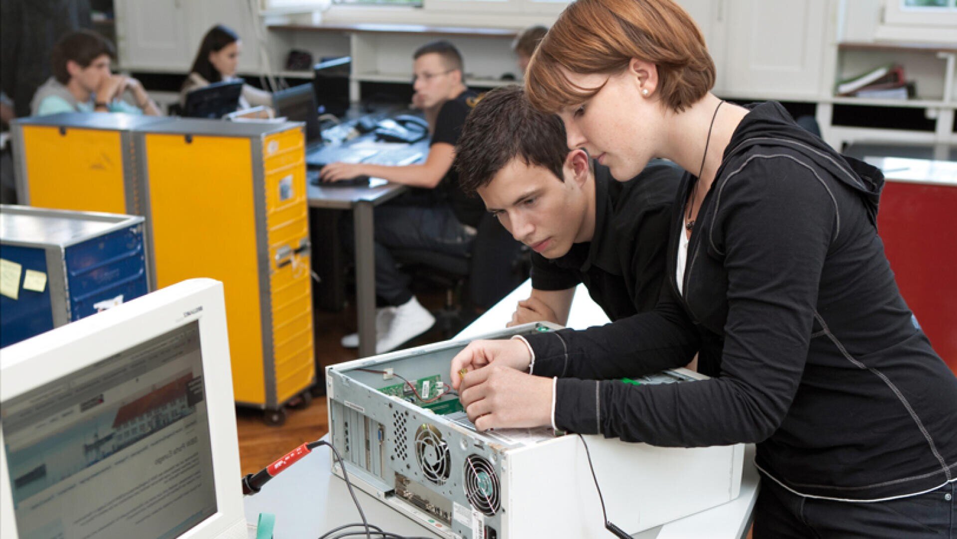 Zwei Schüler arbeiten in einem Klassenzimmer gemeinsam an den inneren Bauteilen eines Desktop-Computers, während andere im Hintergrund arbeiten. Im Vordergrund ist ein Monitor zu sehen, auf dem eine Website angezeigt wird.