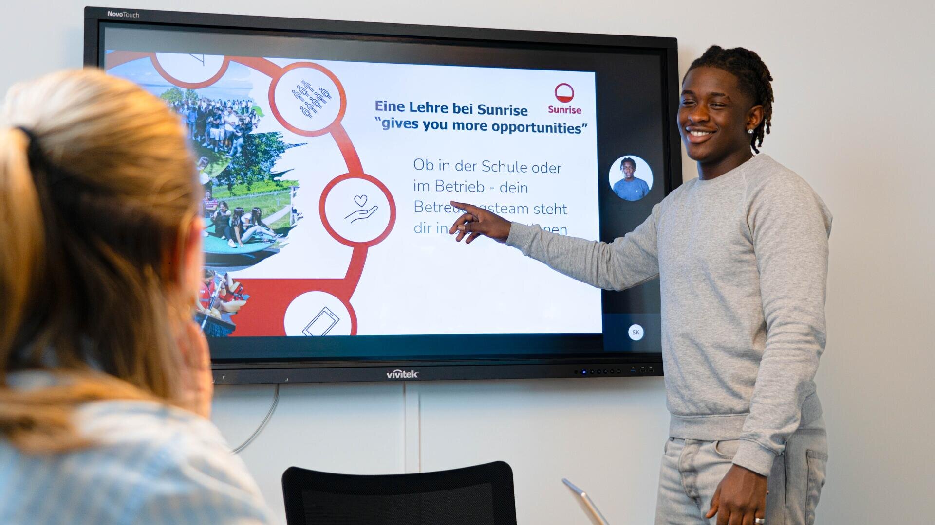 Ein junger Mann steht und zeigt auf einen Präsentationsbildschirm in einem Sitzungszimmer, während eine Frau ihm gegenüber sitzt. Auf dem Bildschirm werden Informationen über die Möglichkeiten bei Sunrise auf Englisch und Deutsch angezeigt.