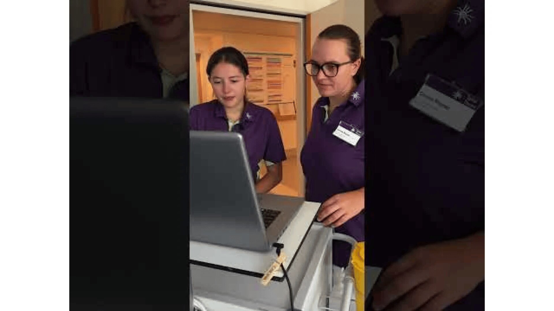 Zwei Mitarbeiter des Gesundheitswesens in lila Uniformen stehen an einem Computerwagen und schauen auf einen Laptop-Bildschirm in einem Krankenhausflur. Einer trägt eine Brille, und beide scheinen auf den Computer konzentriert zu sein.