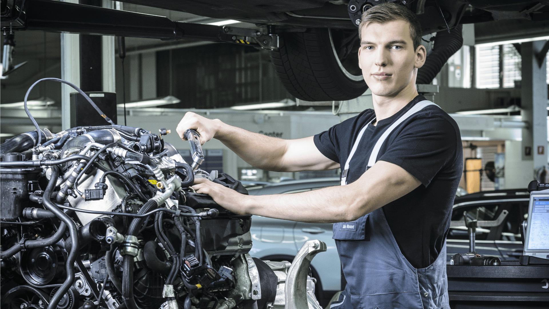 Ein junger männlicher Mechaniker im Overall arbeitet in einer modernen Autowerkstatt mit einem Schraubenschlüssel an einem Automotor und schaut in die Kamera. Ein Auto steht auf einer Hebebühne über ihm, und in der Nähe sind Diagnosegeräte zu sehen.