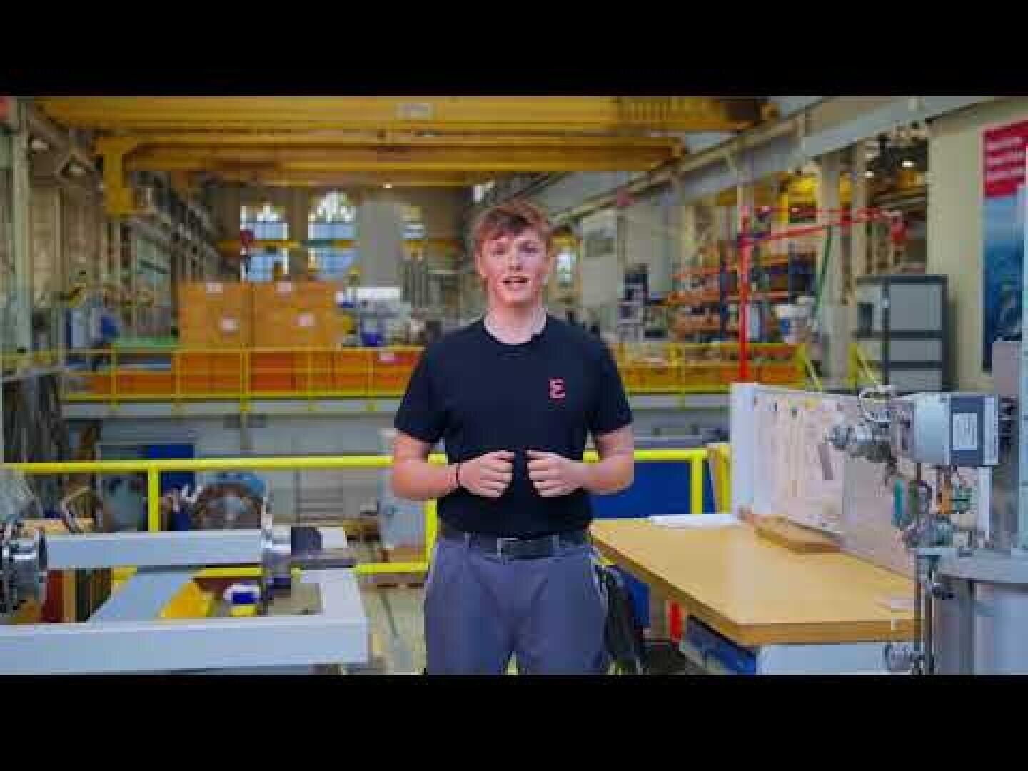 Ein junger Mann steht in einer Industriehalle, trägt ein schwarzes T-Shirt und eine graue Hose, im Hintergrund sind Geräte, Maschinen und gelbe Geländer zu sehen.