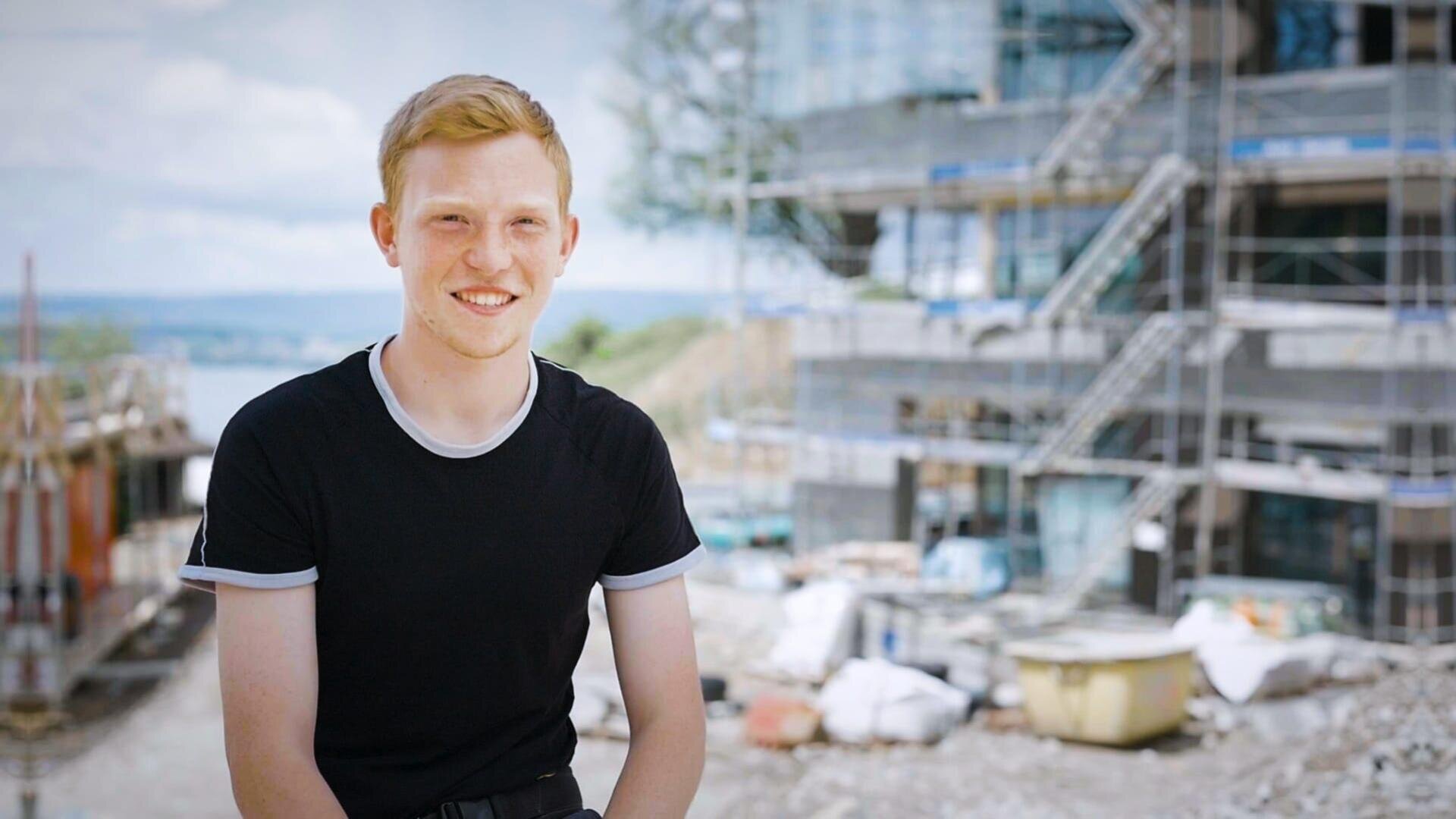 Ein junger Mann mit kurzen blonden Haaren, der ein schwarzes T-Shirt mit weißem Rand trägt, sitzt lächelnd vor einem großen, im Bau befindlichen Gebäude, das von Gerüsten und Baumaterialien umgeben ist.