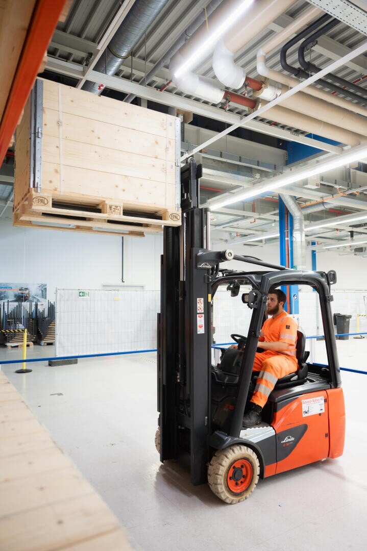 Ein Arbeiter in orangefarbener Uniform bedient einen Gabelstapler und hebt eine große Holzkiste in einem großen, modernen Industrielager.