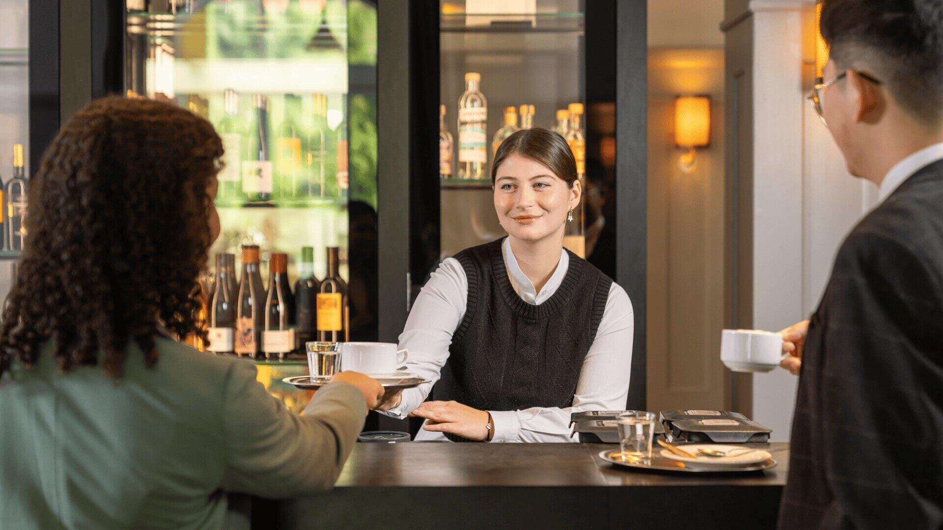 Ein Barkeeper reicht einer sitzenden Frau eine Tasse und eine Untertasse, während ein Mann mit einer Tasse daneben an einer modernen Bar steht, hinter der Flaschen ausgestellt sind.