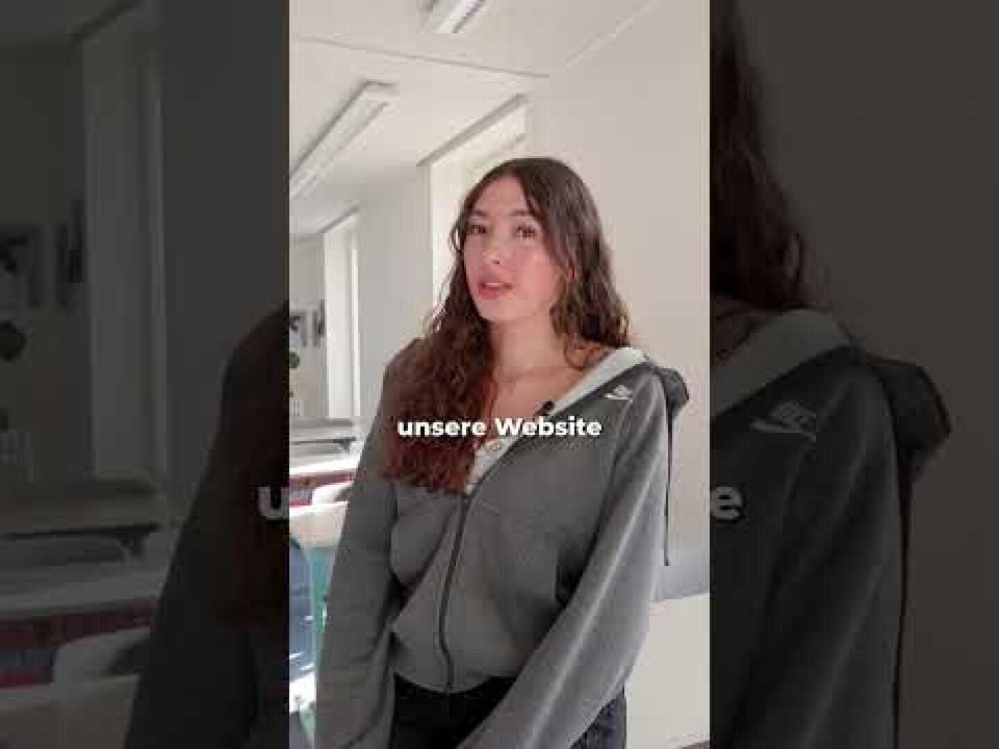 Eine Frau mit langen braunen Haaren, die eine graue Jacke mit Reißverschluss trägt, steht in einem Raum. In der Bildmitte ist der Text „unsere Website“ zu sehen.