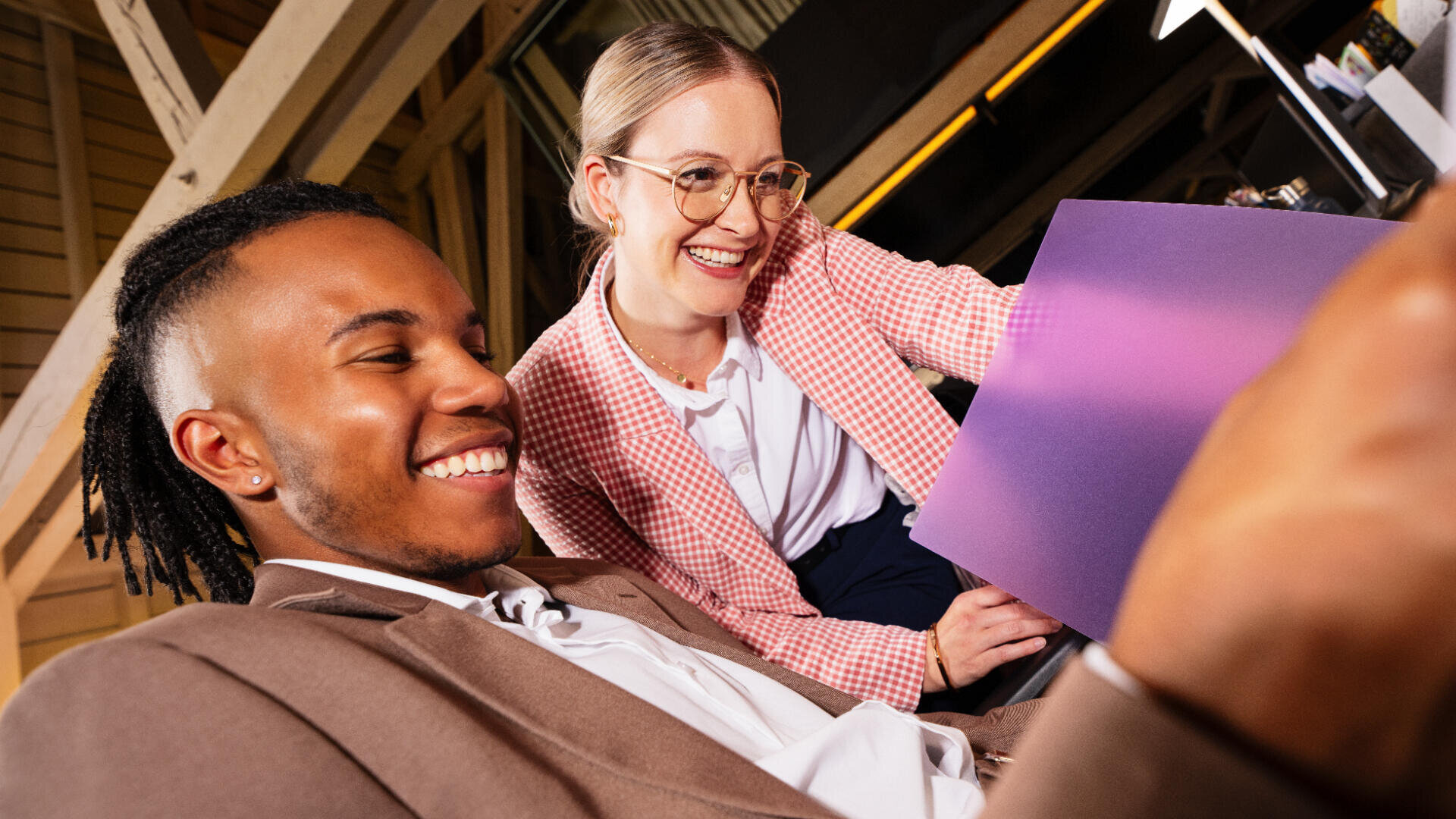 Zwei Personen in Geschäftskleidung lächeln und schauen gemeinsam auf ein Tablet. Der Vordermann hat kurze Dreadlocks und trägt einen hellbraunen Blazer, der Hintermann trägt eine Brille und einen rosa karierten Blazer und hält einen lila Ordner.