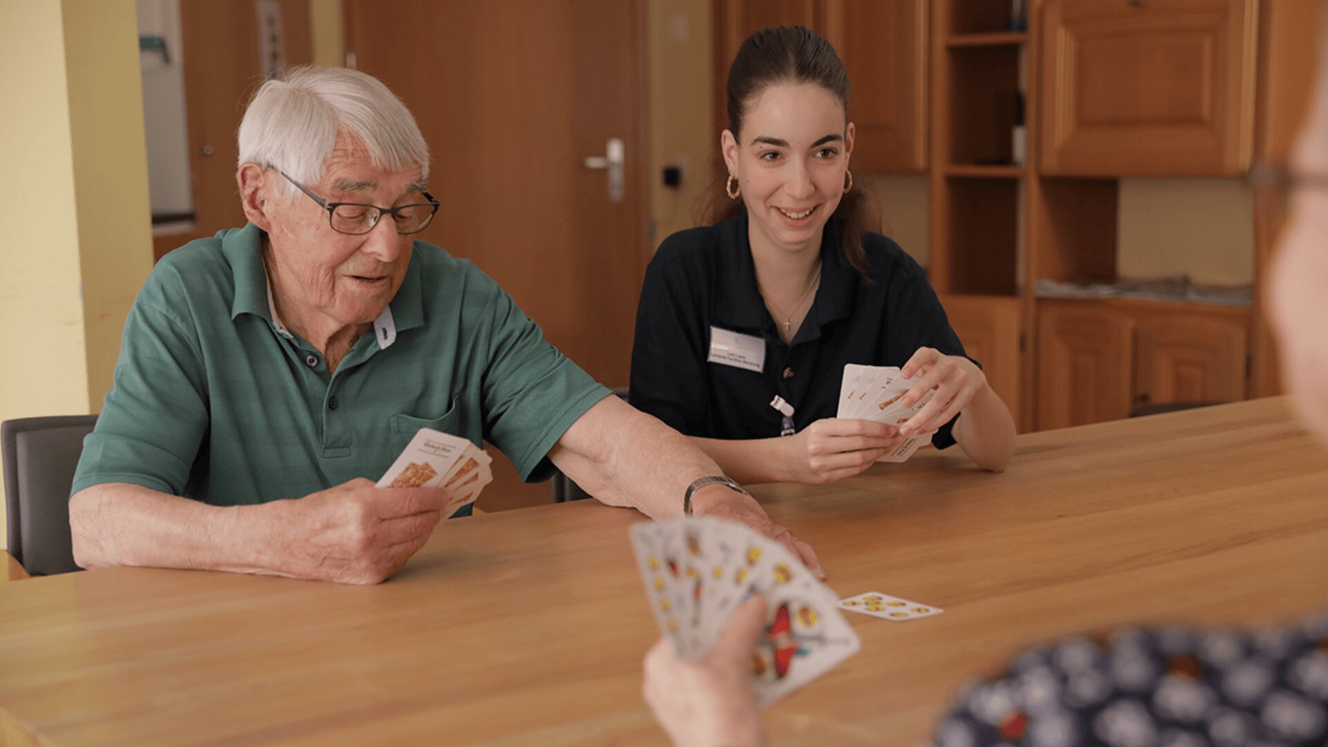 Ein älterer Mann und eine junge Frau sitzen an einem Holztisch und spielen lächelnd Karten mit einer anderen Person, die mit dem Rücken zur Kamera steht.