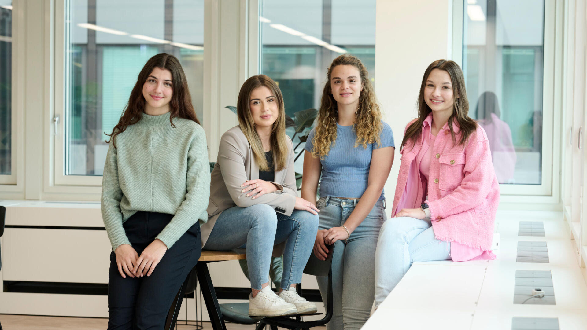 Vier junge Frauen sitzen und posieren zusammen in einem Innenraum vor großen Fenstern. Sie sind leger gekleidet und lächeln, während im Hintergrund eine helle und moderne Büroumgebung zu sehen ist.