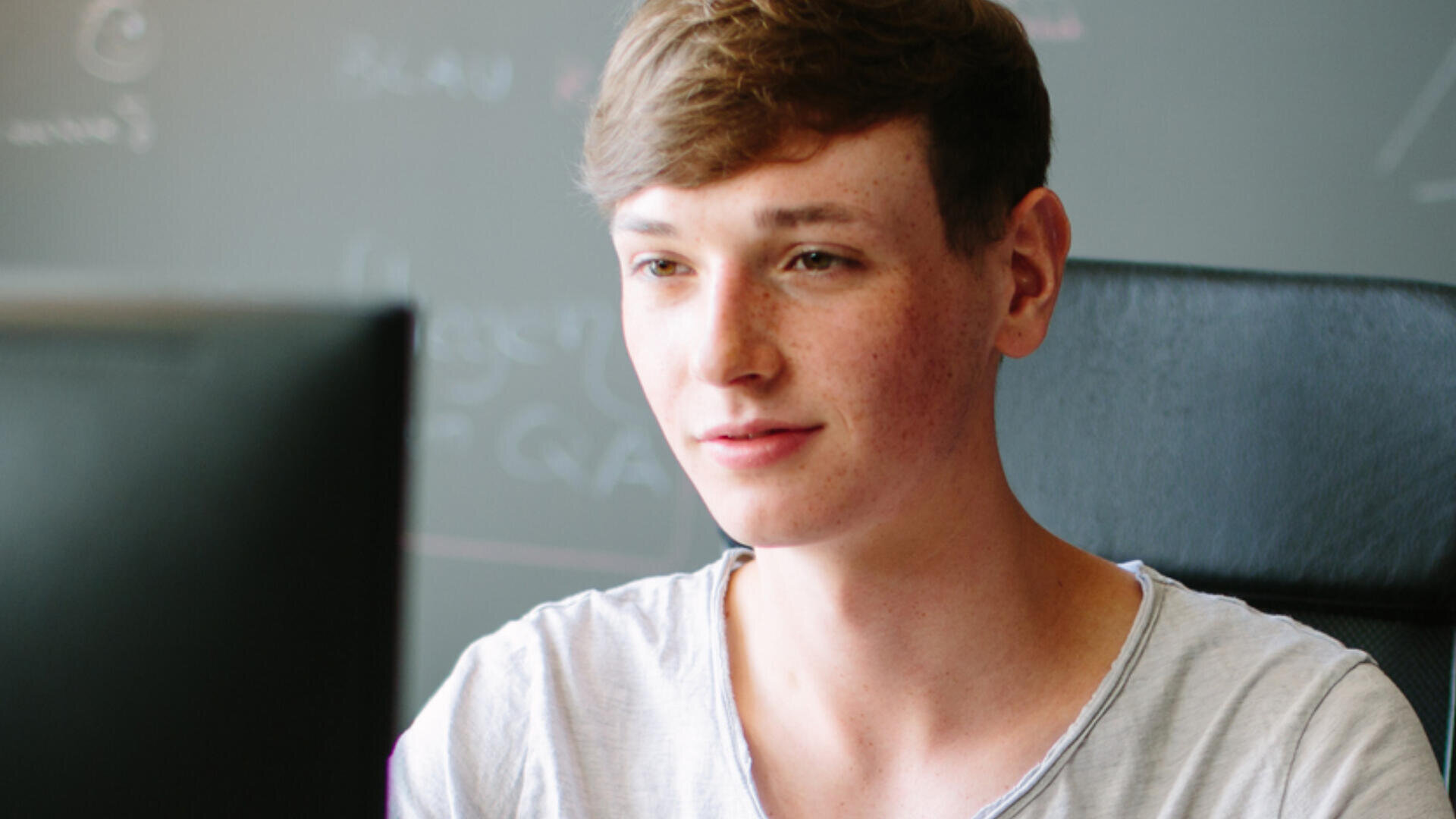 Eine junge Person mit kurzen braunen Haaren und einem hellen Hemd sitzt an einem Schreibtisch und schaut auf einen Computerbildschirm, im Hintergrund ist eine verschwommene Kreidetafel zu sehen.