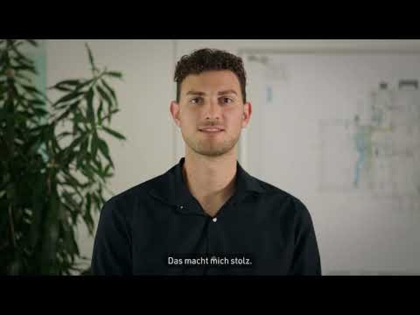 Ein junger Mann mit kurzen lockigen Haaren und einem dunklen Hemd steht in einem Raum neben einer hohen Pflanze. An der Wand hinter ihm hängt eine Blaupause. Der deutsche Text am unteren Rand lautet: "Das macht mich stolz.