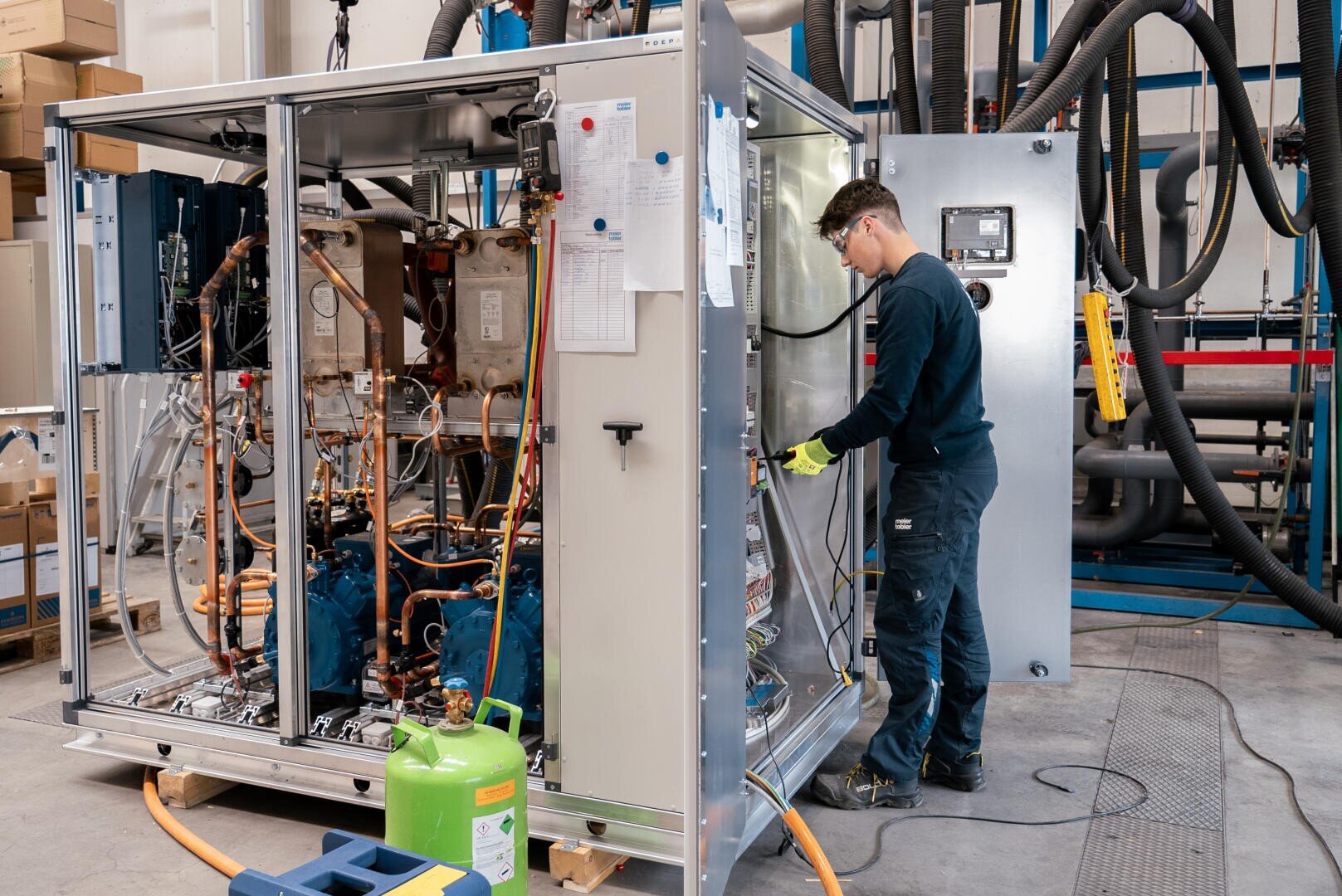 Ein Techniker mit Schutzhandschuhen arbeitet in einer Werkstatt an einem offenen industriellen Schaltschrank mit freiliegenden Kabeln und Komponenten. Um den Arbeitsbereich herum sind aufgerollte Kabel, Werkzeuge und Geräte zu sehen.