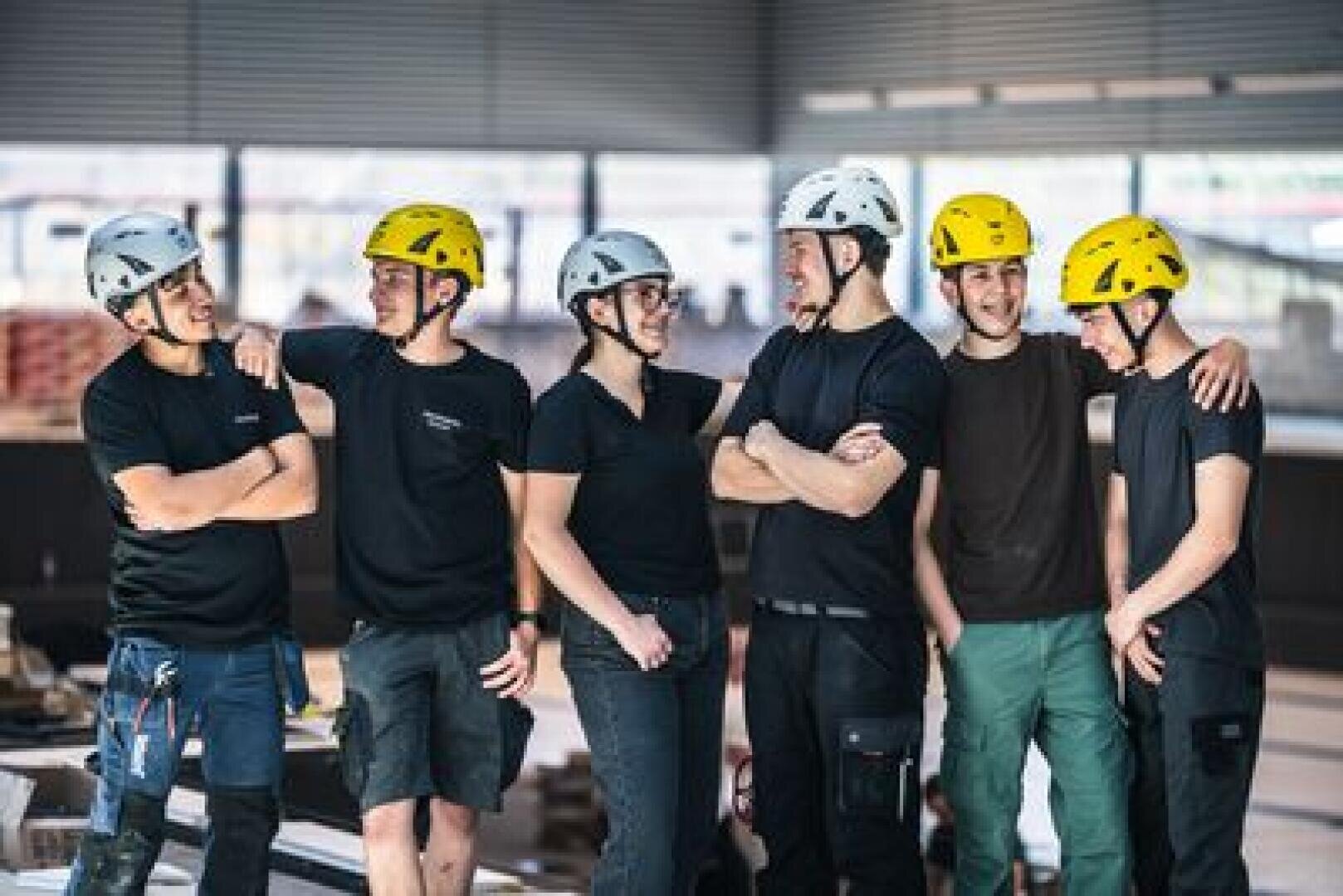Sechs Personen, die Schutzhelme und legere Arbeitskleidung tragen, stehen in einem Raum, lächeln und unterhalten sich. Der Hintergrund lässt auf eine Baustelle oder eine Industrieumgebung schließen.