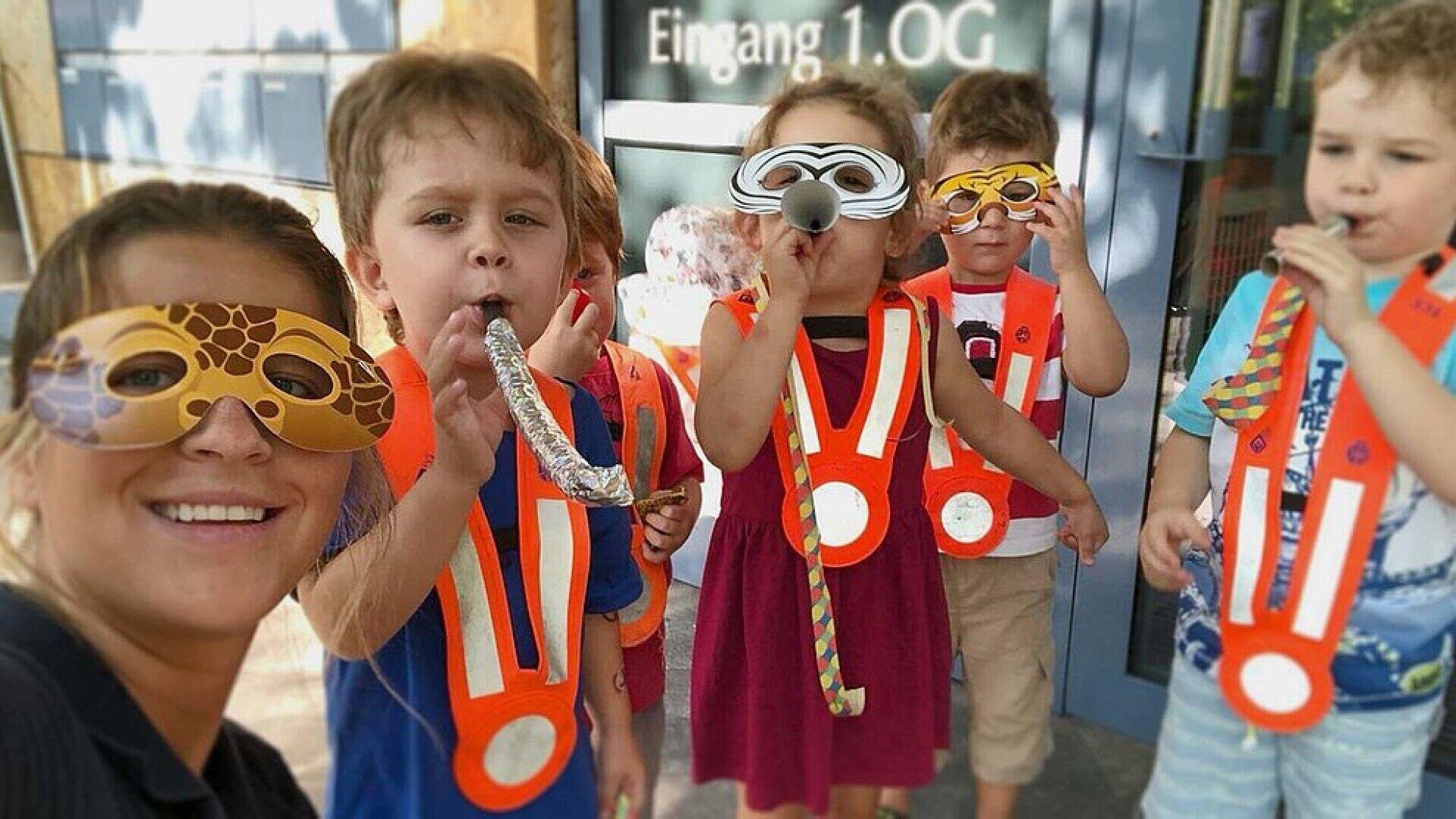 Eine Frau und fünf kleine Kinder mit festlichen Masken und orangefarbenen Sicherheitswesten feiern gemeinsam, blasen in Partyhupen und lächeln vor einem Gebäude mit Glastüren mit der Aufschrift Eingang 1. OG.