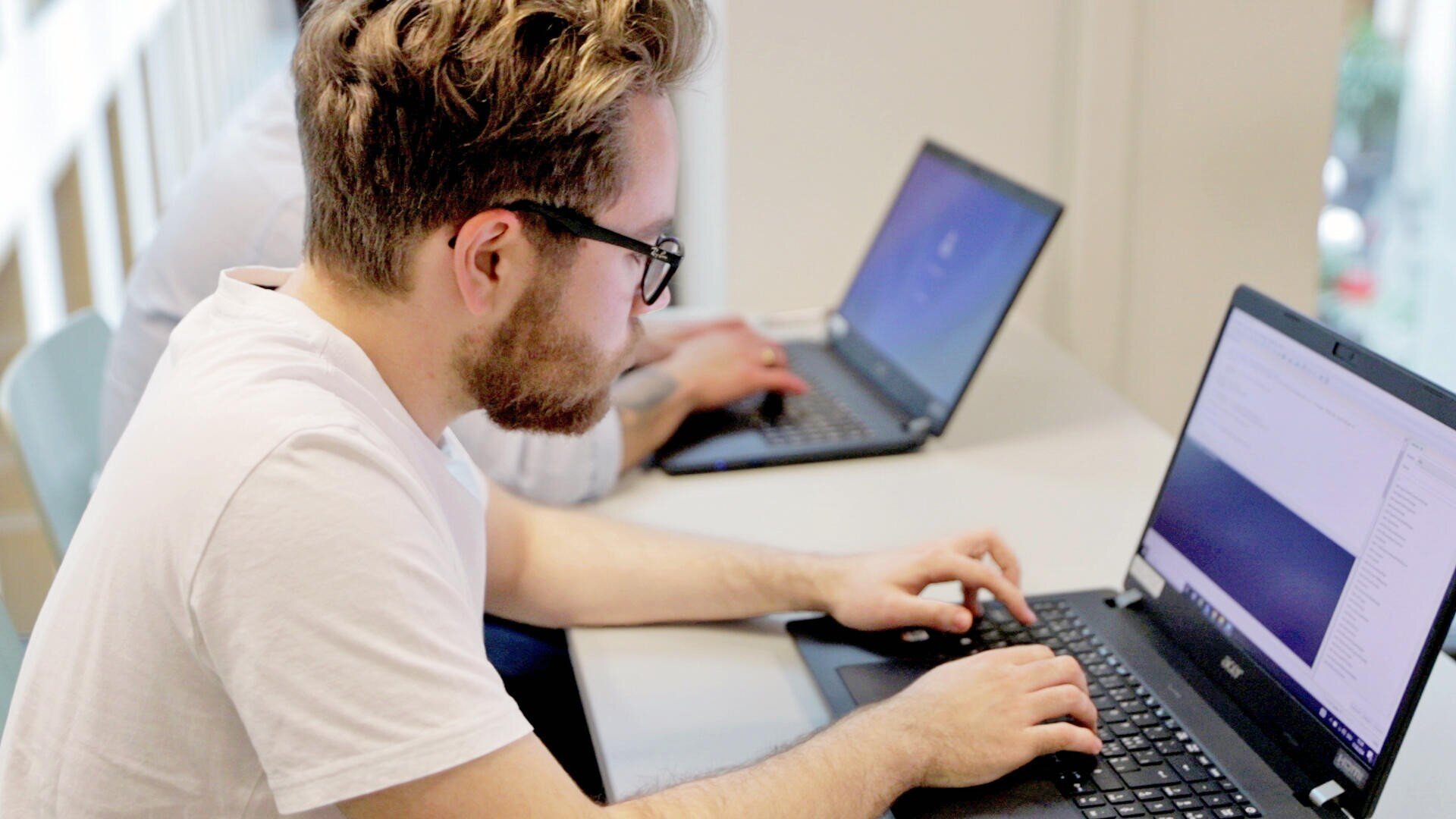 Ein Mann mit Brille und Bart tippt an einem Laptop an einem Schreibtisch, während eine andere Person im Hintergrund ebenfalls an einem Laptop arbeitet. Beide wirken konzentriert in einem hellen, modernen Arbeitsbereich.