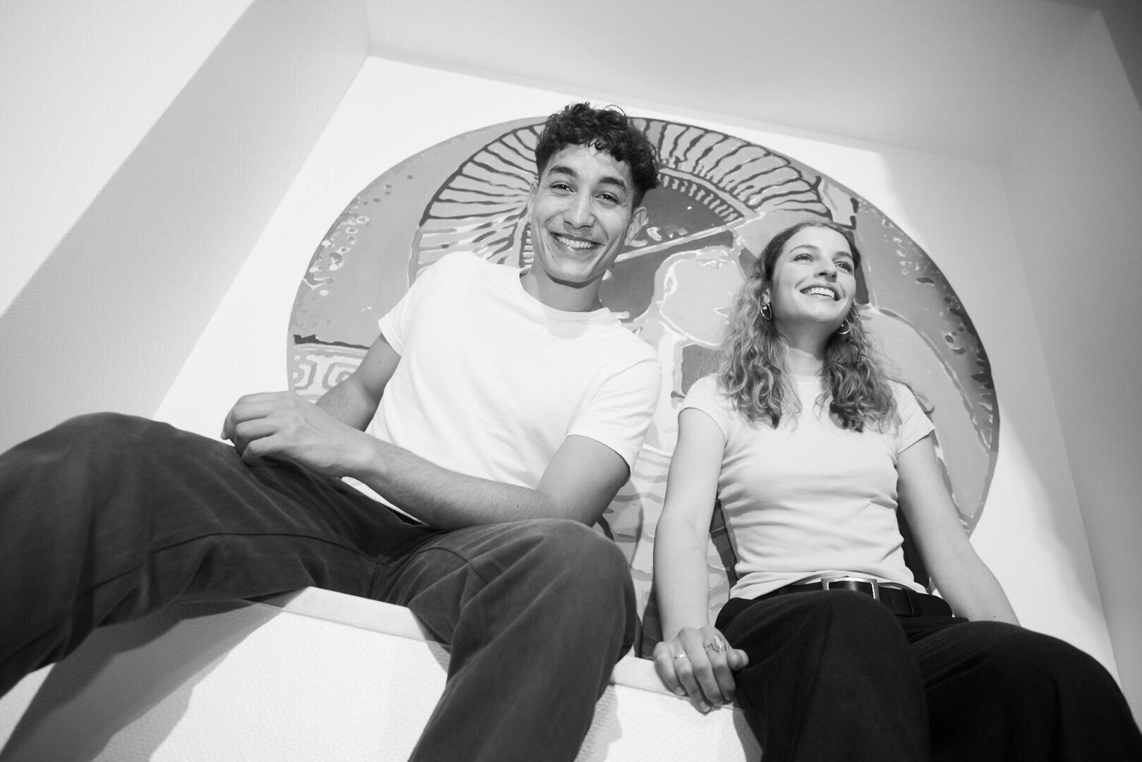 Ein junger Mann und eine junge Frau sitzen nebeneinander, halten sich an den Händen und lächeln. Sie tragen Freizeitkleidung und sitzen vor einem großen, runden Kunstwerk an der Wand. Das Foto ist schwarz-weiß und wurde aus einer niedrigen Perspektive aufgenommen.