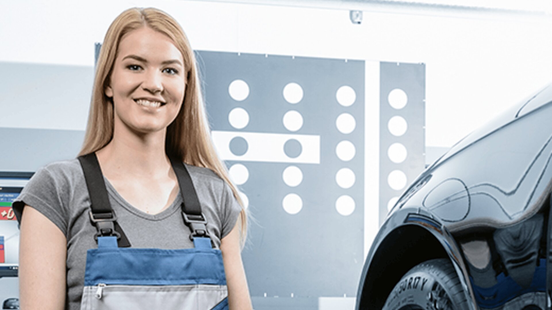 Eine lächelnde Frau in einem Mechaniker-Overall steht neben einem Fahrzeug in einer Garage oder Werkstatt, wobei im Hintergrund Kfz-Ausrüstung und eine Kalibrierungsanzeige zu sehen sind.