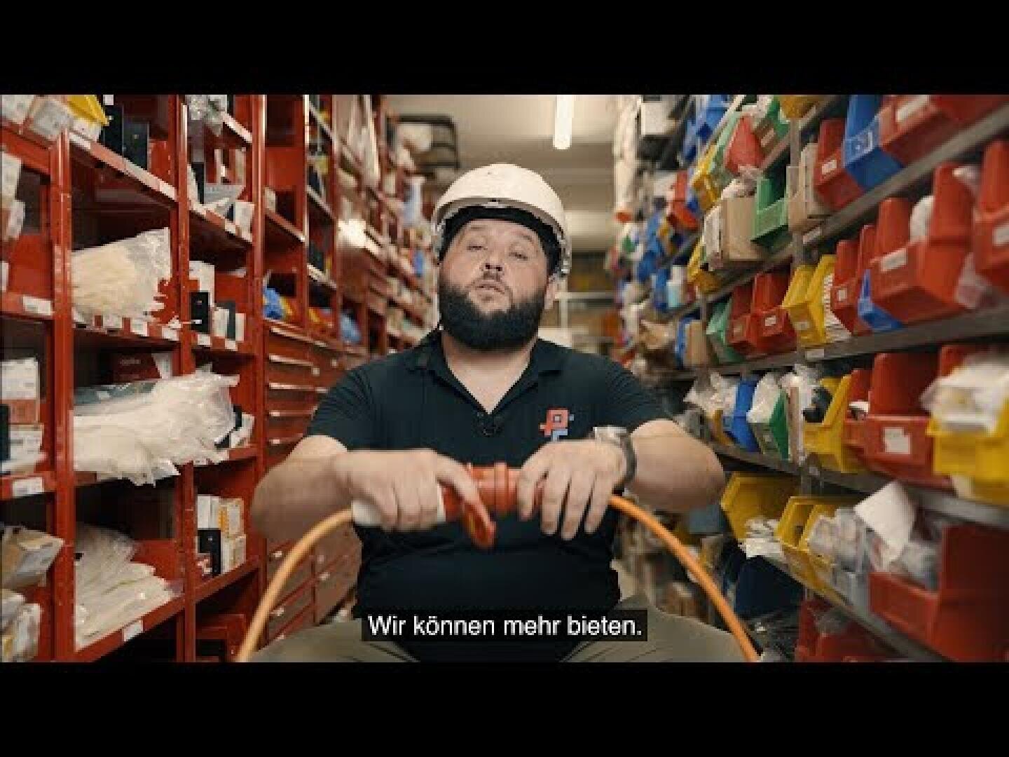 Ein Mann mit einem Schutzhelm sitzt in einem Lagergang mit bunten Behältern und hält ein Kabel in der Hand. Er trägt ein schwarzes T-Shirt mit einem Logo. In den deutschen Untertiteln steht: „Wir können mehr bieten…“.