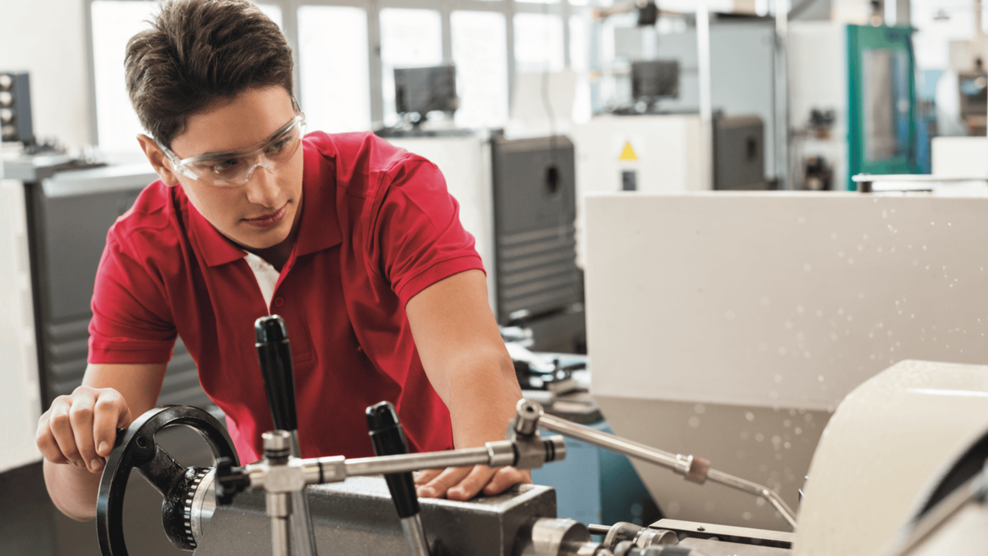 Ein junger Mensch mit Schutzbrille und rotem Hemd bedient eine Drehmaschine in einer modernen Industriehalle.