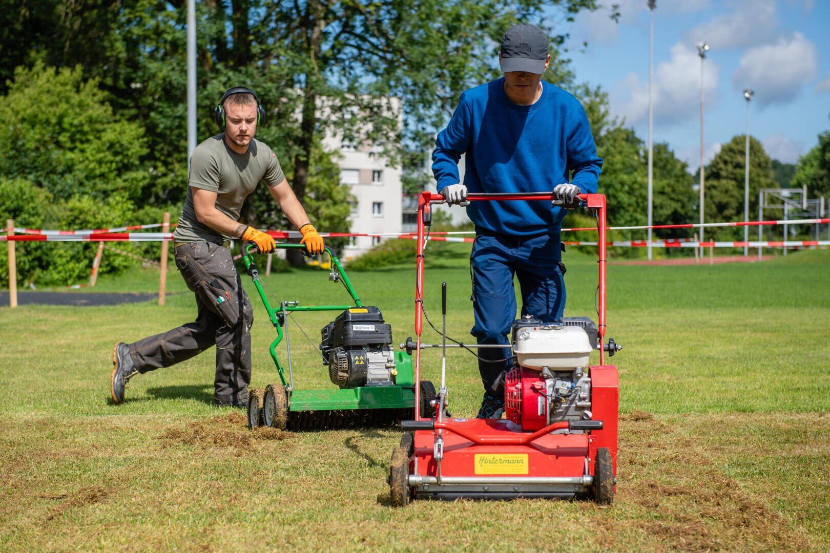 Zwei Personen benutzen motorisierte Rasengeräte auf einer Wiese. Eine Person in einem blauen Outfit schiebt eine rote Maschine, während die andere, in einem grünen Hemd, eine grüne Maschine bedient. Im Hintergrund sind Bäume und Gebäude zu sehen.