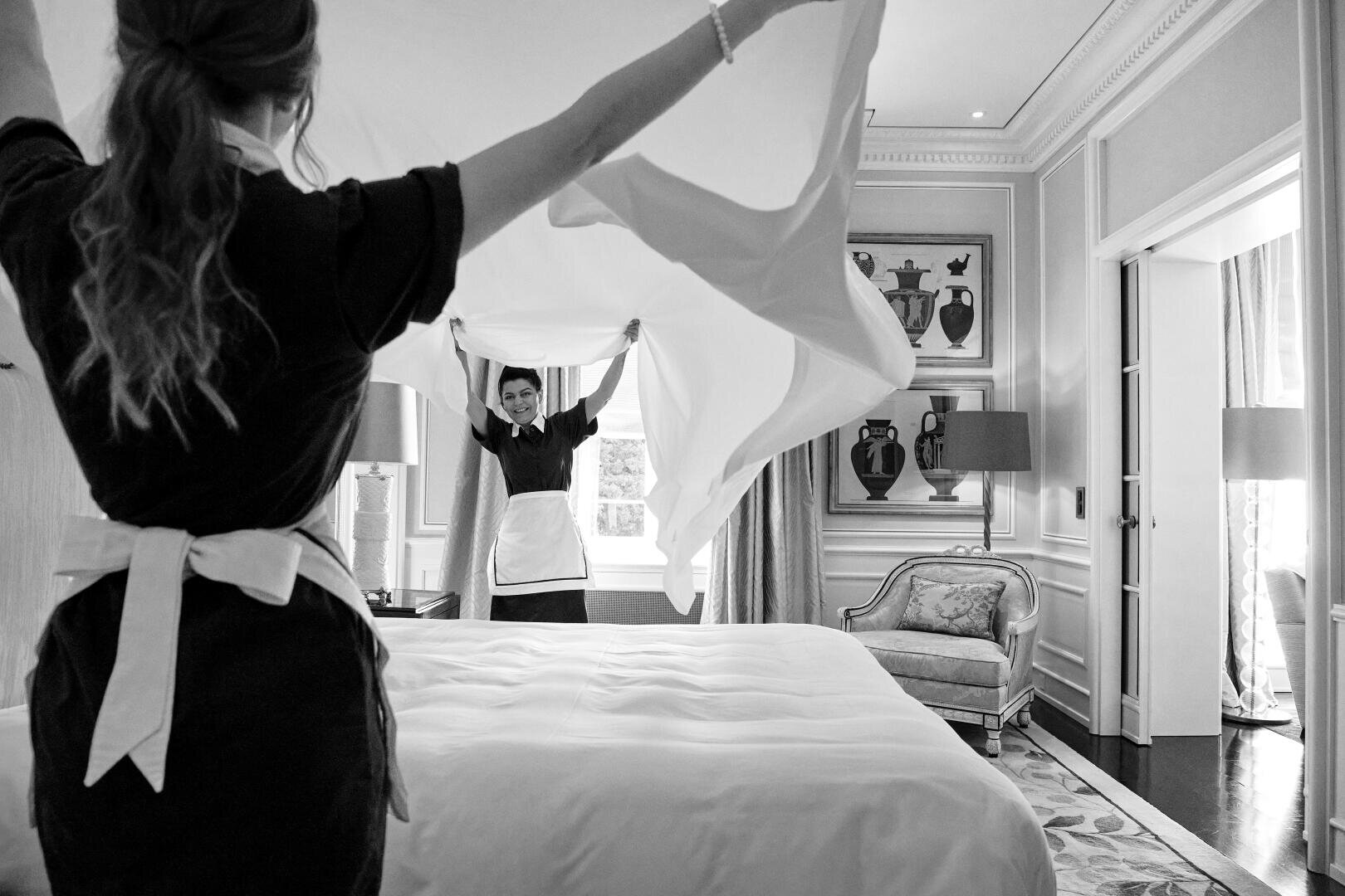 Zwei Hotelzimmermädchen in Uniform machen ein Bett in einem eleganten, gut beleuchteten Hotelzimmer, indem sie gemeinsam ein Laken anheben und ausbreiten. Zur Ausstattung gehören gerahmte Kunstwerke, ein großes Fenster und Plüschmöbel.